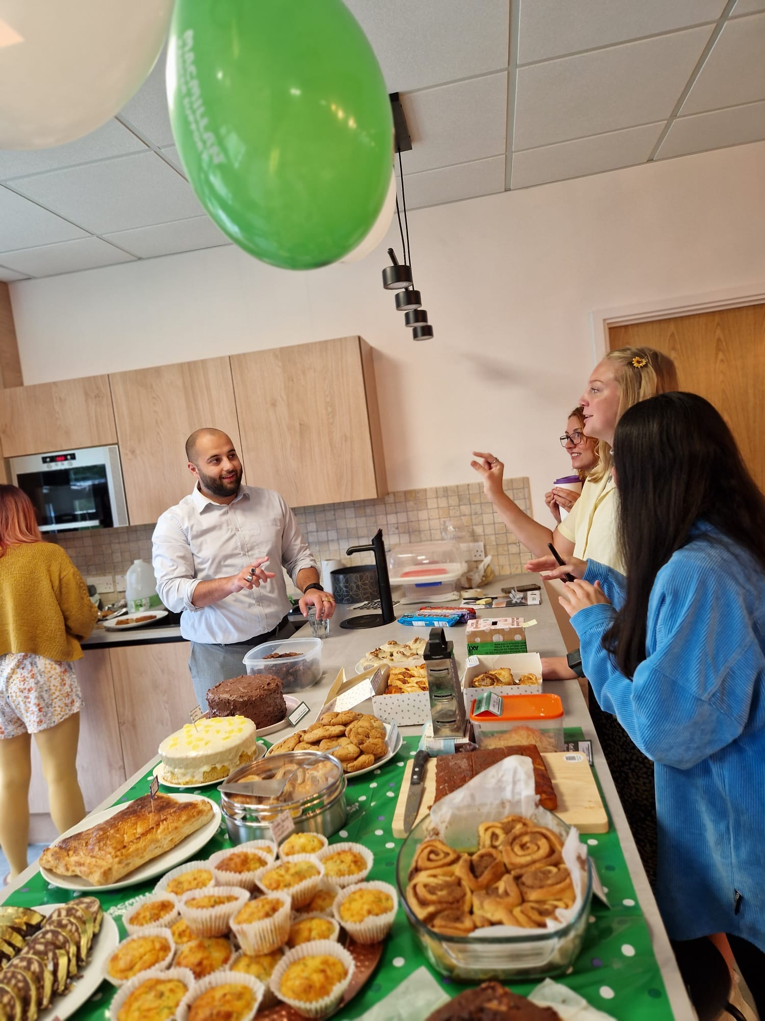People in modern kitchen with food spread on green polka dot tablecloth, green balloon hanging from ceiling, wooden cabinets.