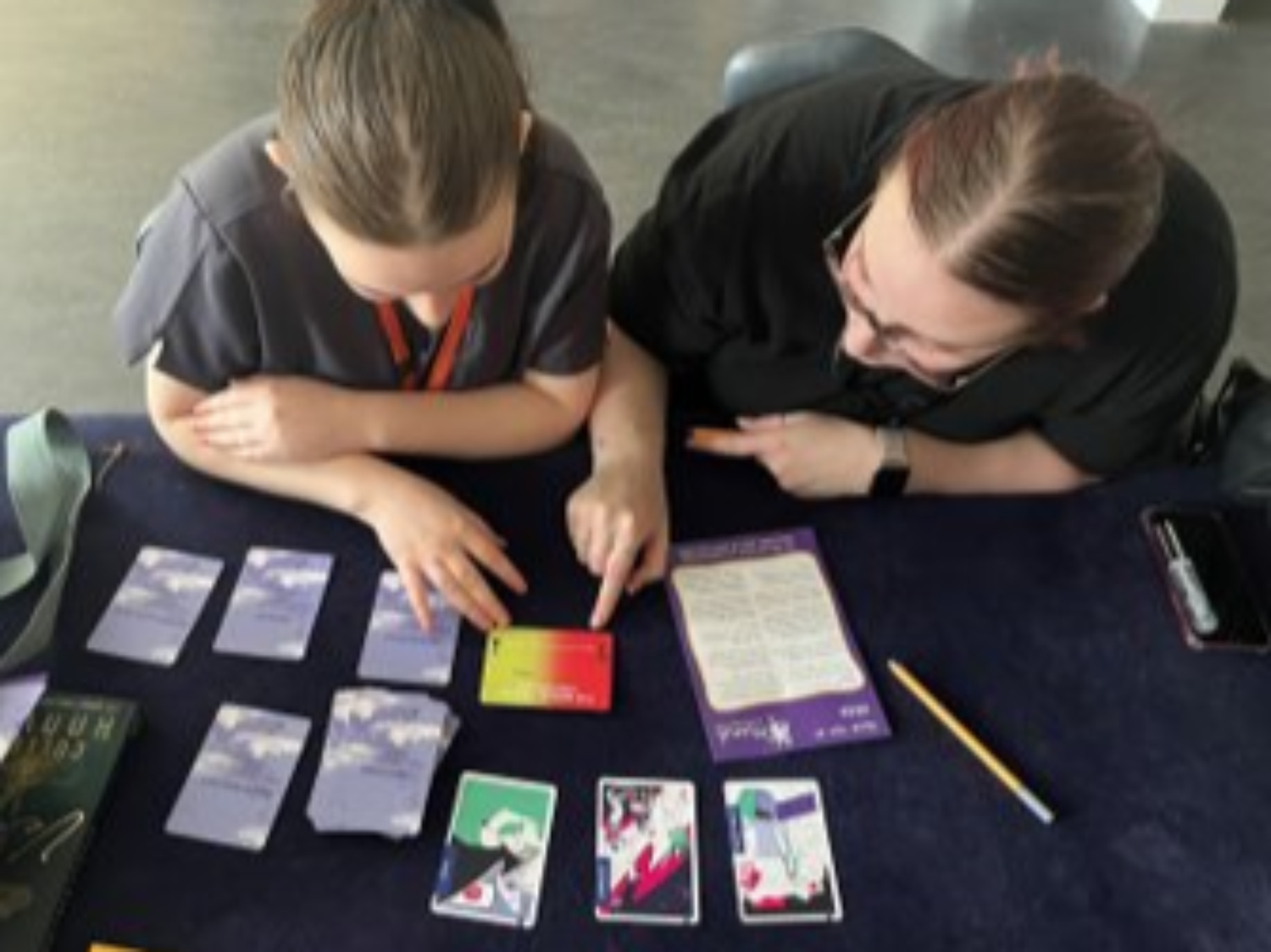 Two people in dark clothing examining colourful cards arranged on a dark surface, with papers and a pen visible nearby.