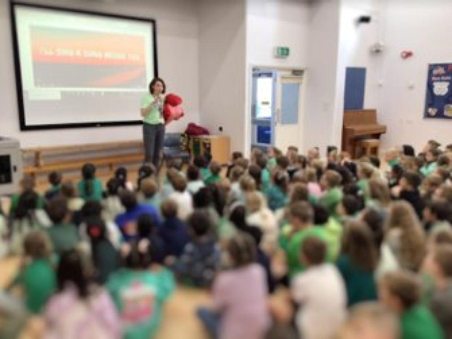 Woman speaking to seated children in green school uniforms in school hall with projector screen and wooden stage.