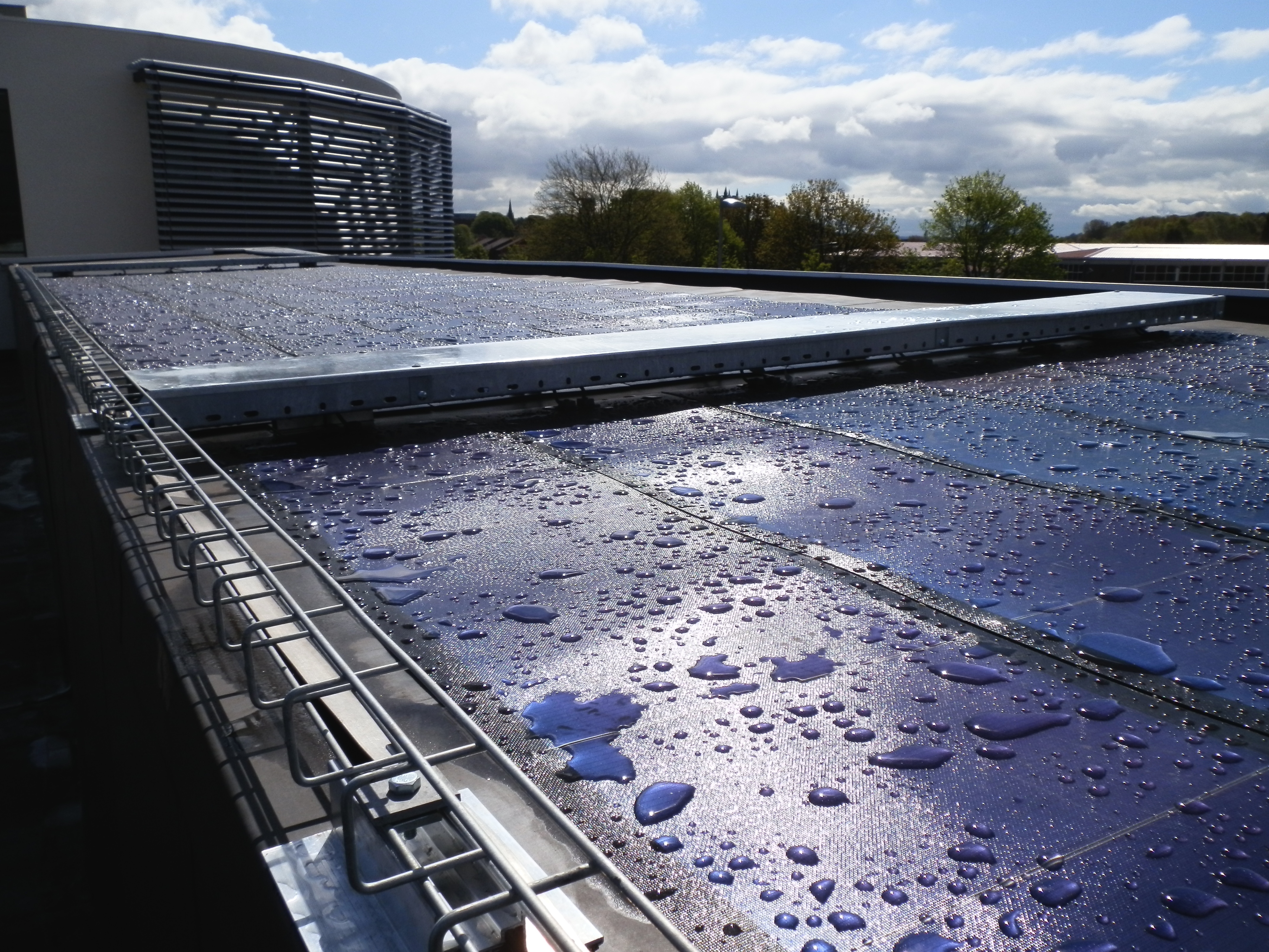 Wet rooftop with water puddles reflecting cloudy sky, metal drainage system, modern building and trees in background.
