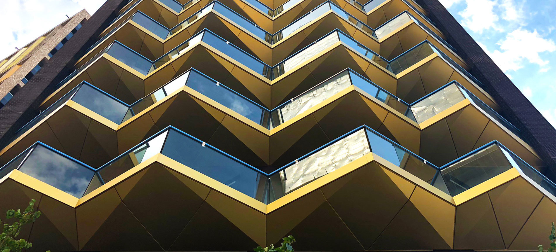 Modern building with zigzag golden balconies and blue glass windows creating geometric pattern against blue sky with white clouds.