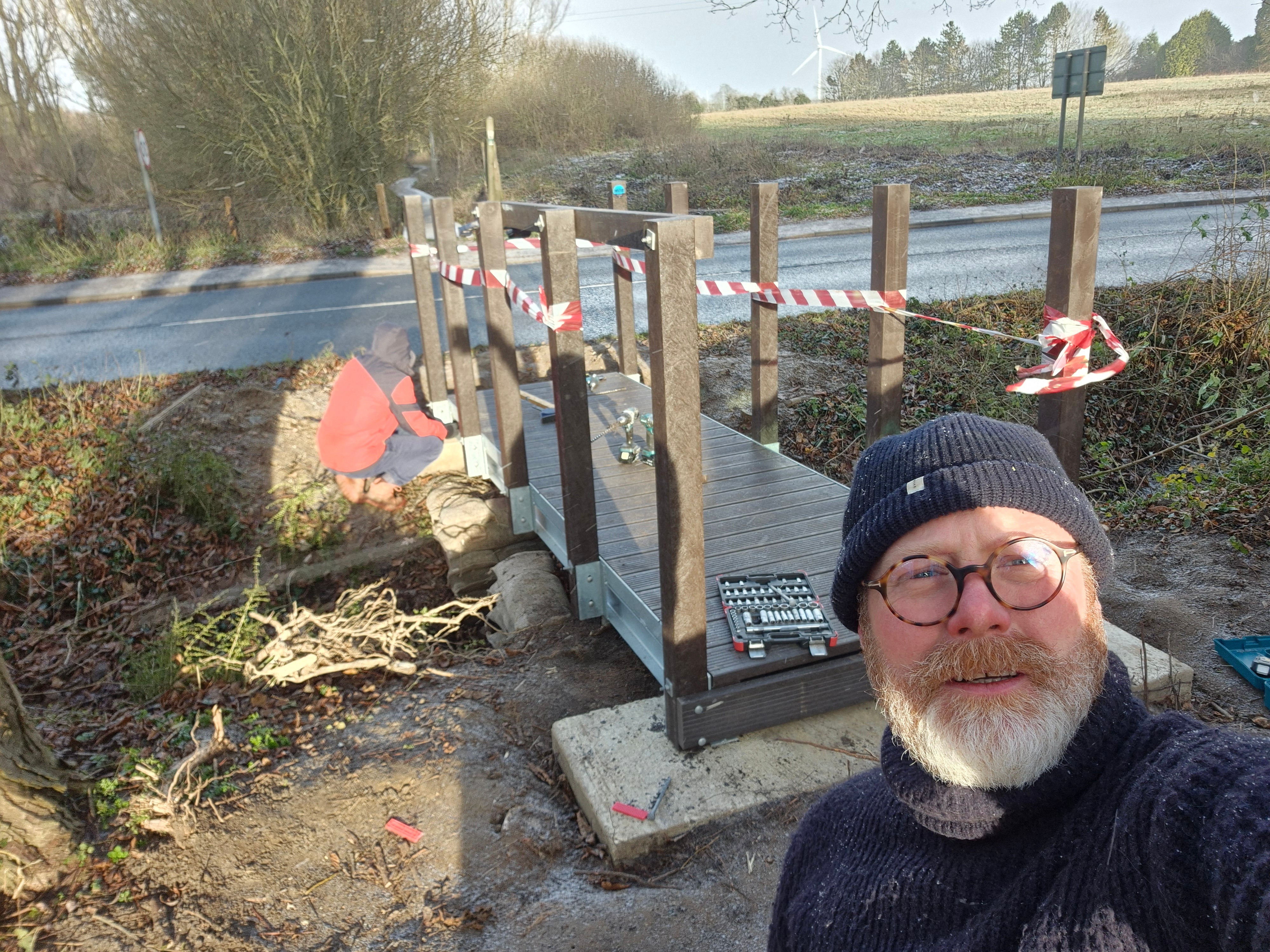 Bearded man in glasses and beanie taking selfie beside wooden footbridge under construction with metal grating and safety tape.