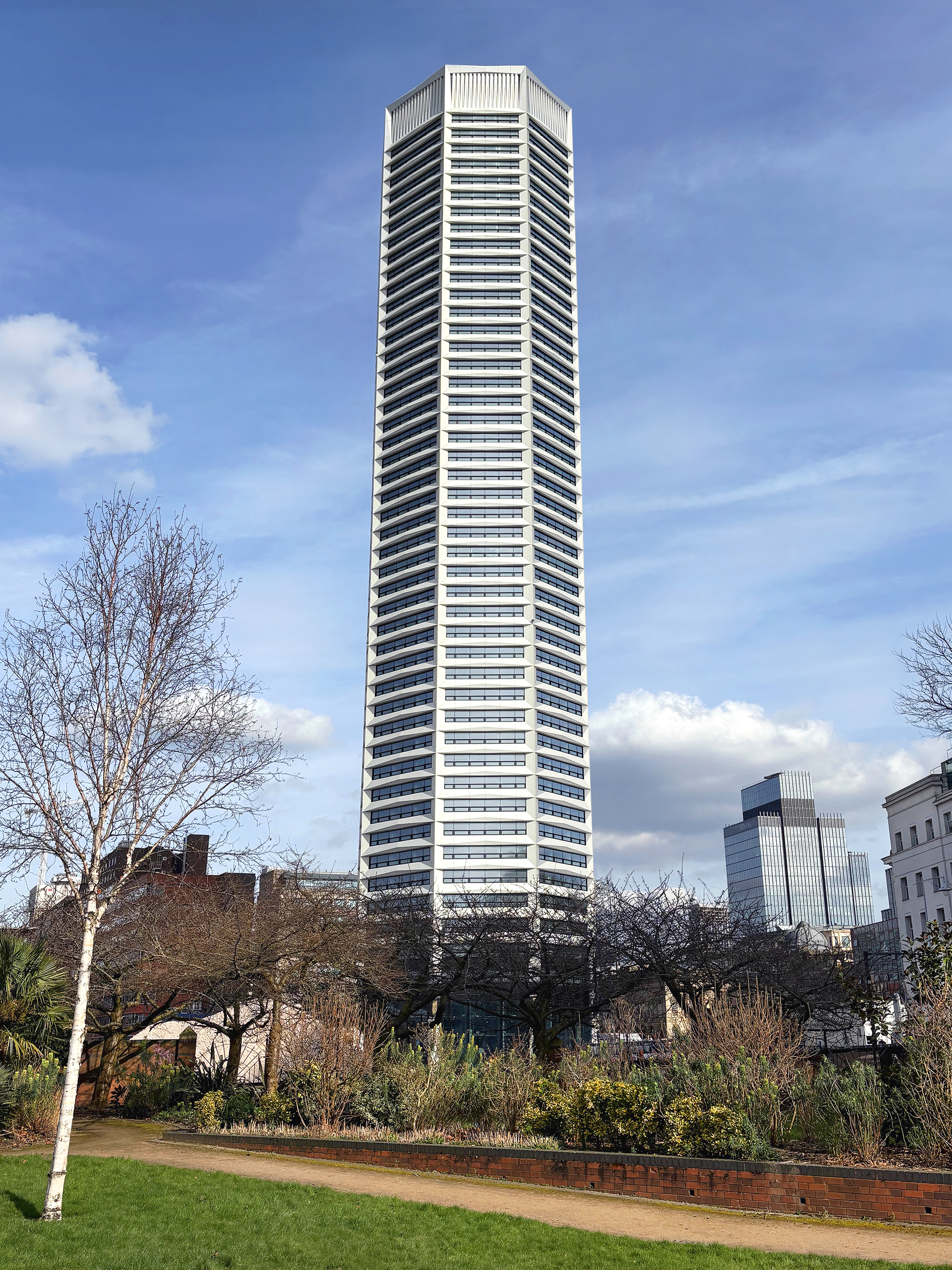 Tall white and grey striped tower block against blue sky with white clouds, viewed from park with green grass and bare trees.