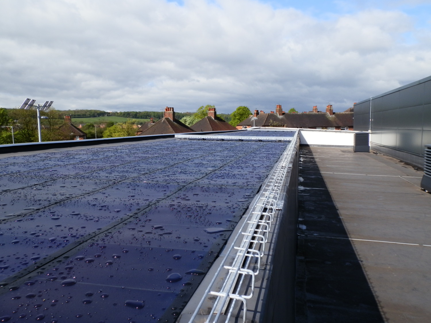 Dark blue solar panels on flat rooftop with metal safety railing, residential houses visible in background under cloudy sky.