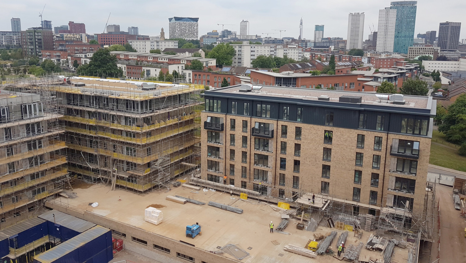 Aerial view of two modern residential buildings under construction with scaffolding, surrounded by urban cityscape and trees.