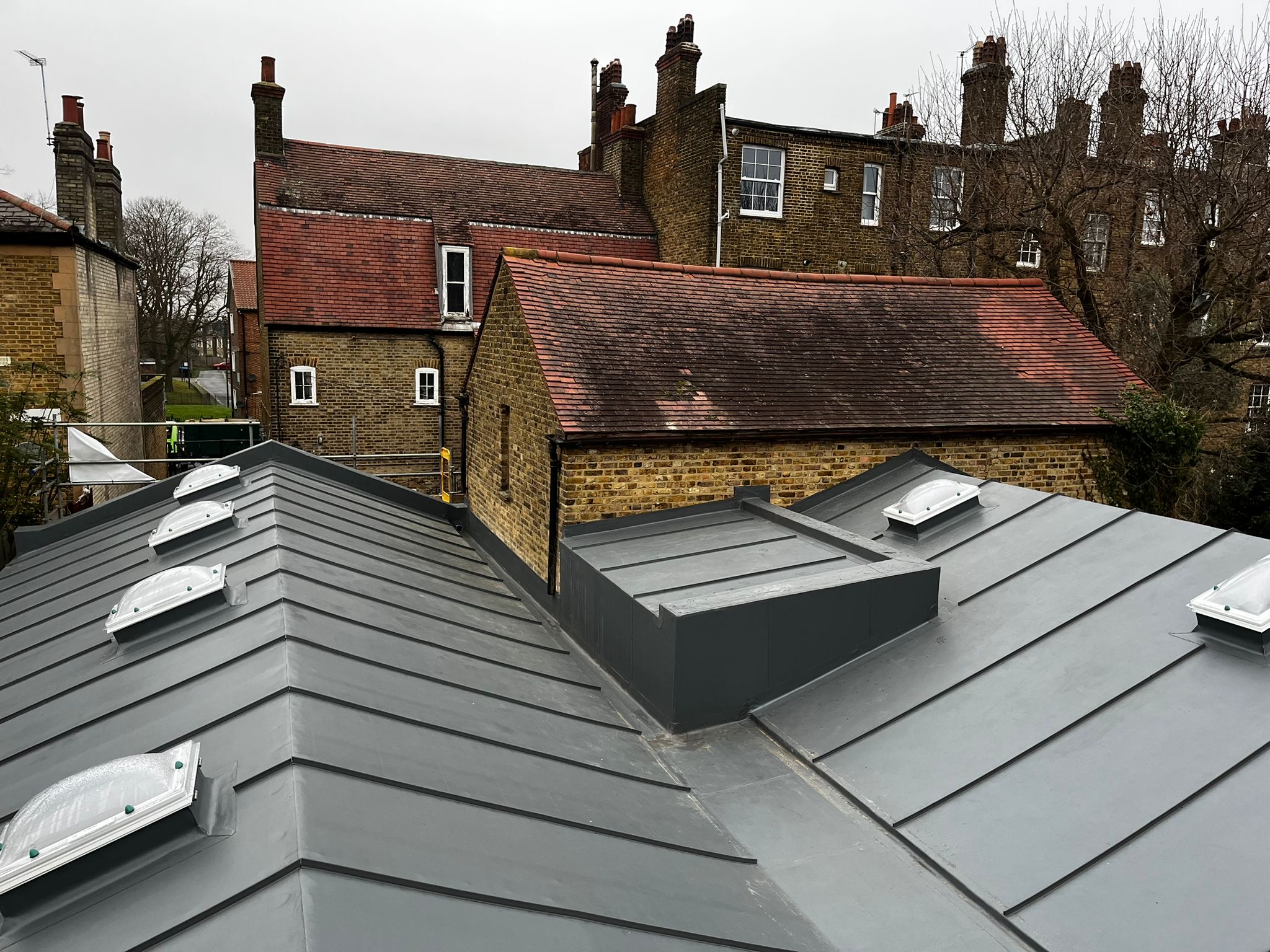Grey metal roofing in foreground with traditional British terraced houses behind featuring brick walls, chimney stacks and tiled roofs.