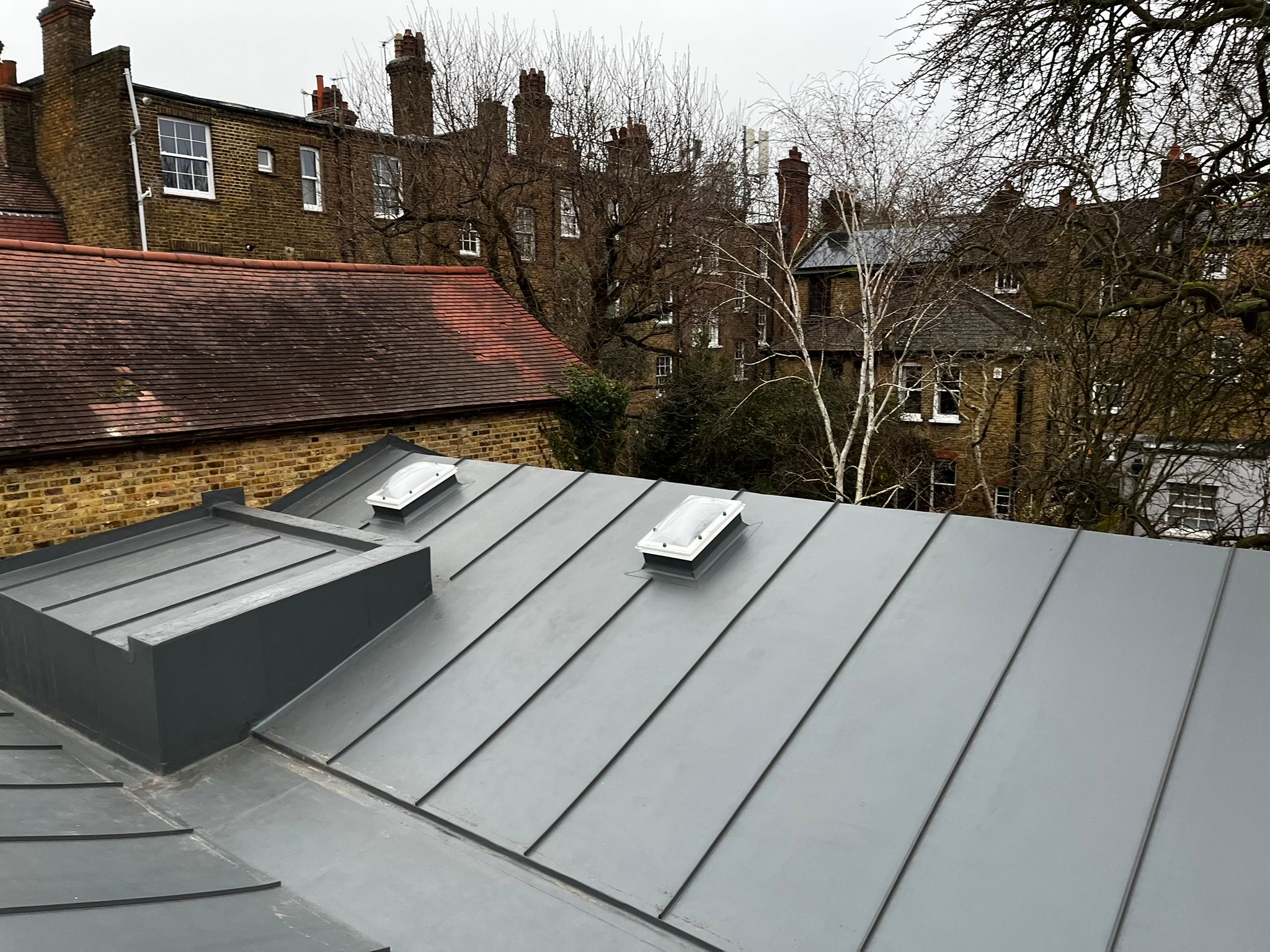 Grey metal roof with standing seam panels and roof lights, overlooking Victorian terraced houses with red brick and chimneys.