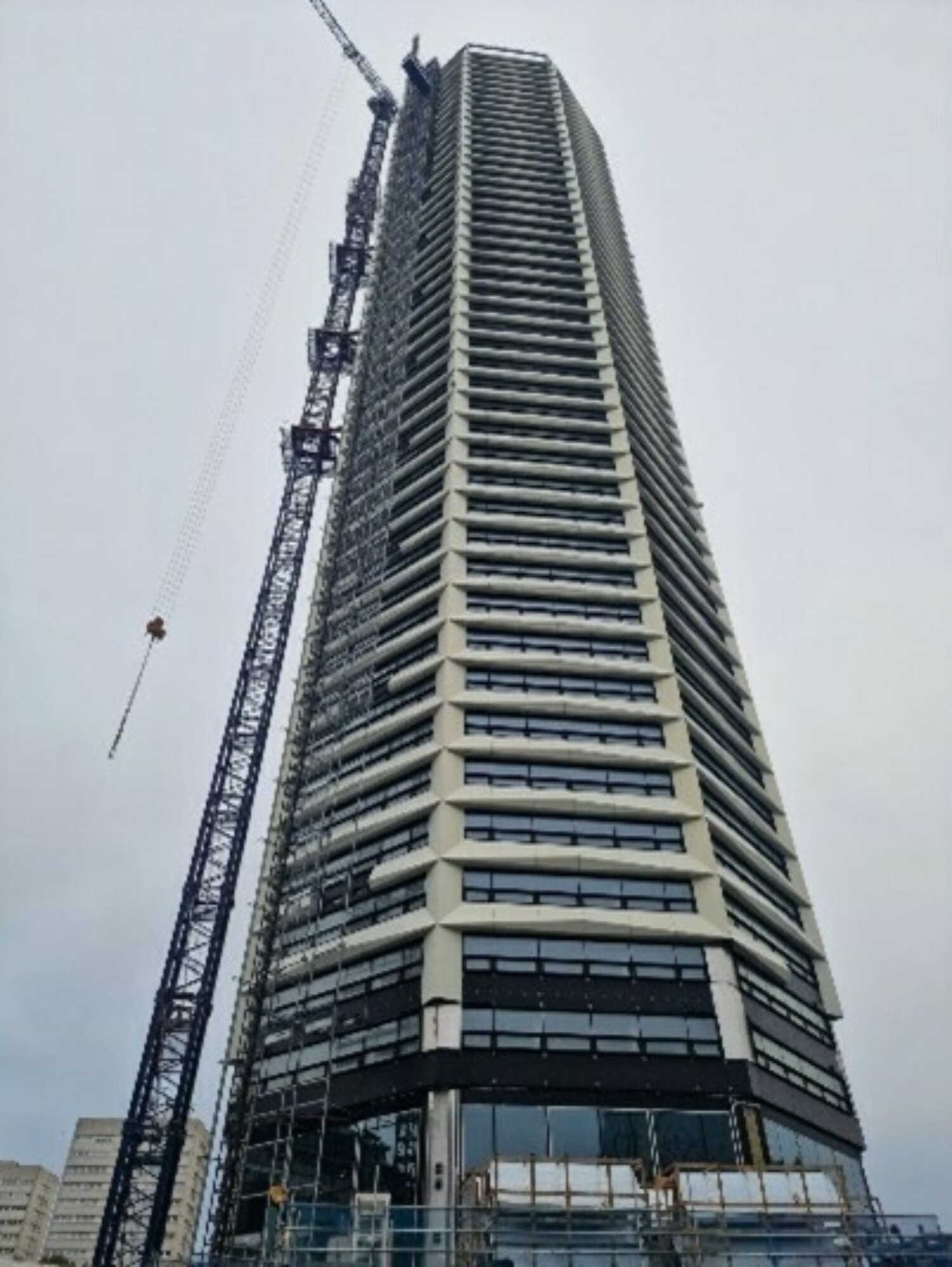 Tall residential tower with glass balconies and white structural elements, construction crane visible, overcast sky background.