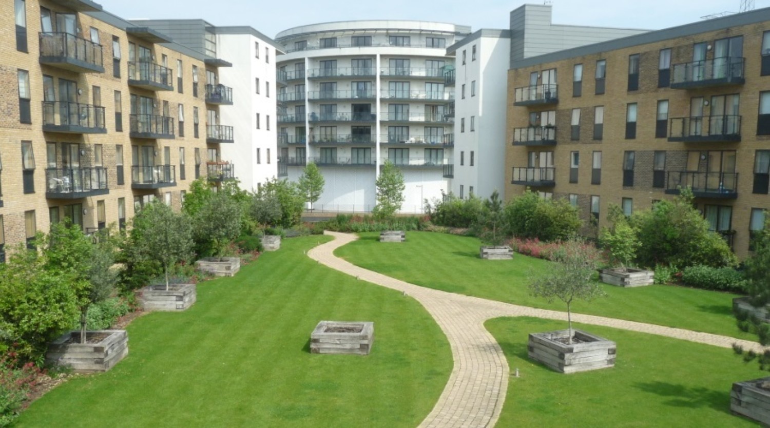 Modern residential complex with brick and white buildings surrounding central courtyard featuring curved pathways, green lawn, and raised planters.