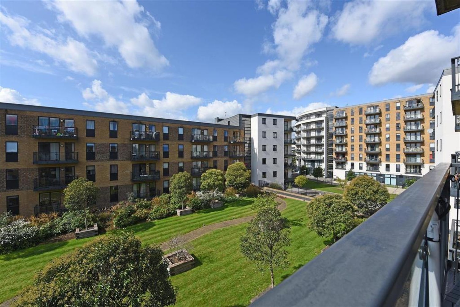 Modern apartment blocks surrounding central green courtyard with lawn and trees, viewed from balcony under blue sky with white clouds.