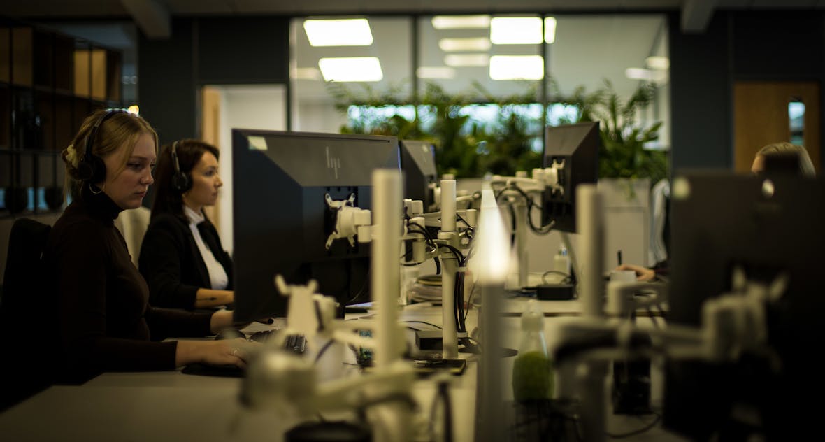 Two people working at computer workstations in dimly lit office with glass partitions and plants visible in background.