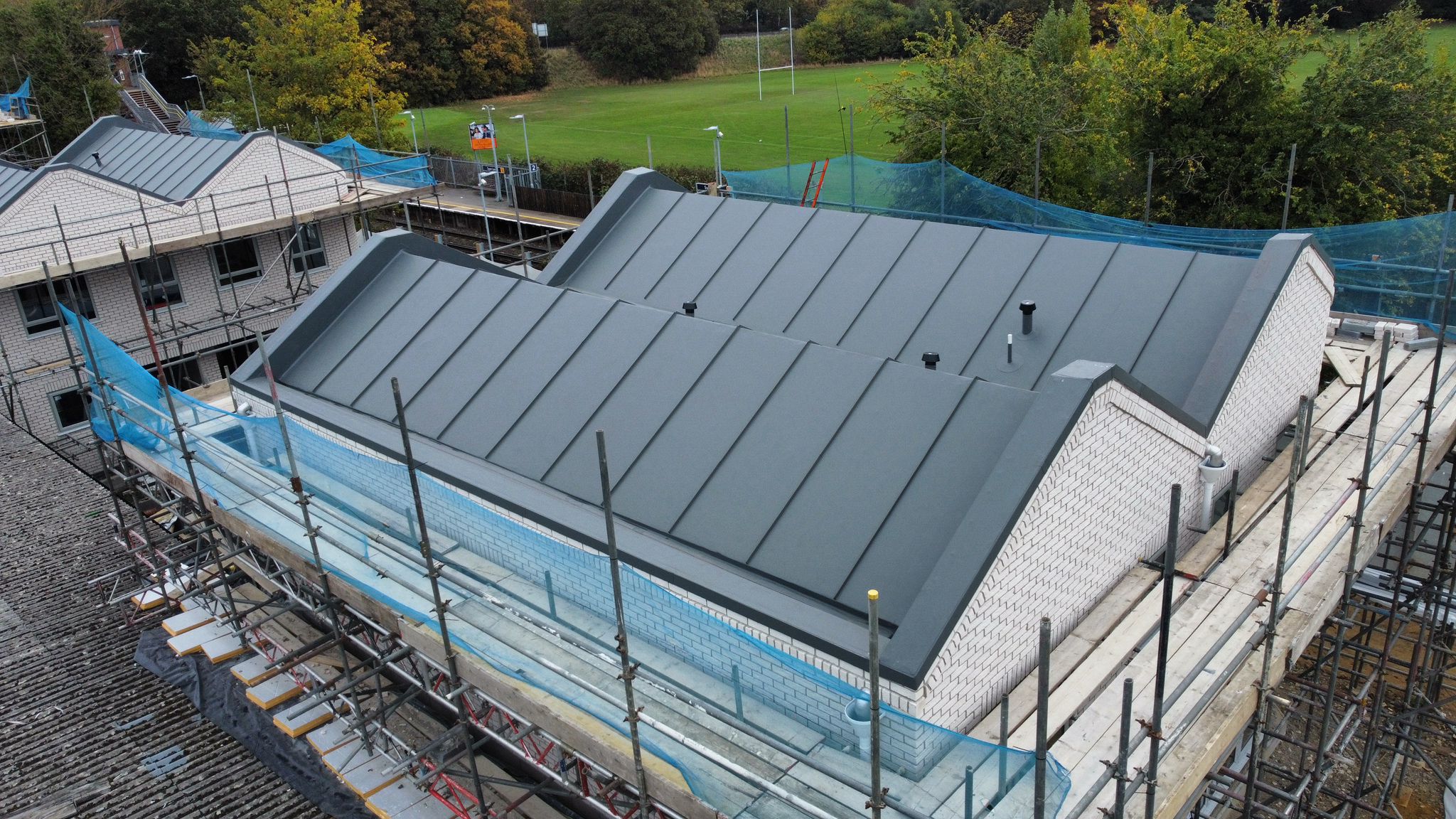 Aerial view of house under construction with grey metal roof, blue protective sheeting, scaffolding, and green fields beyond