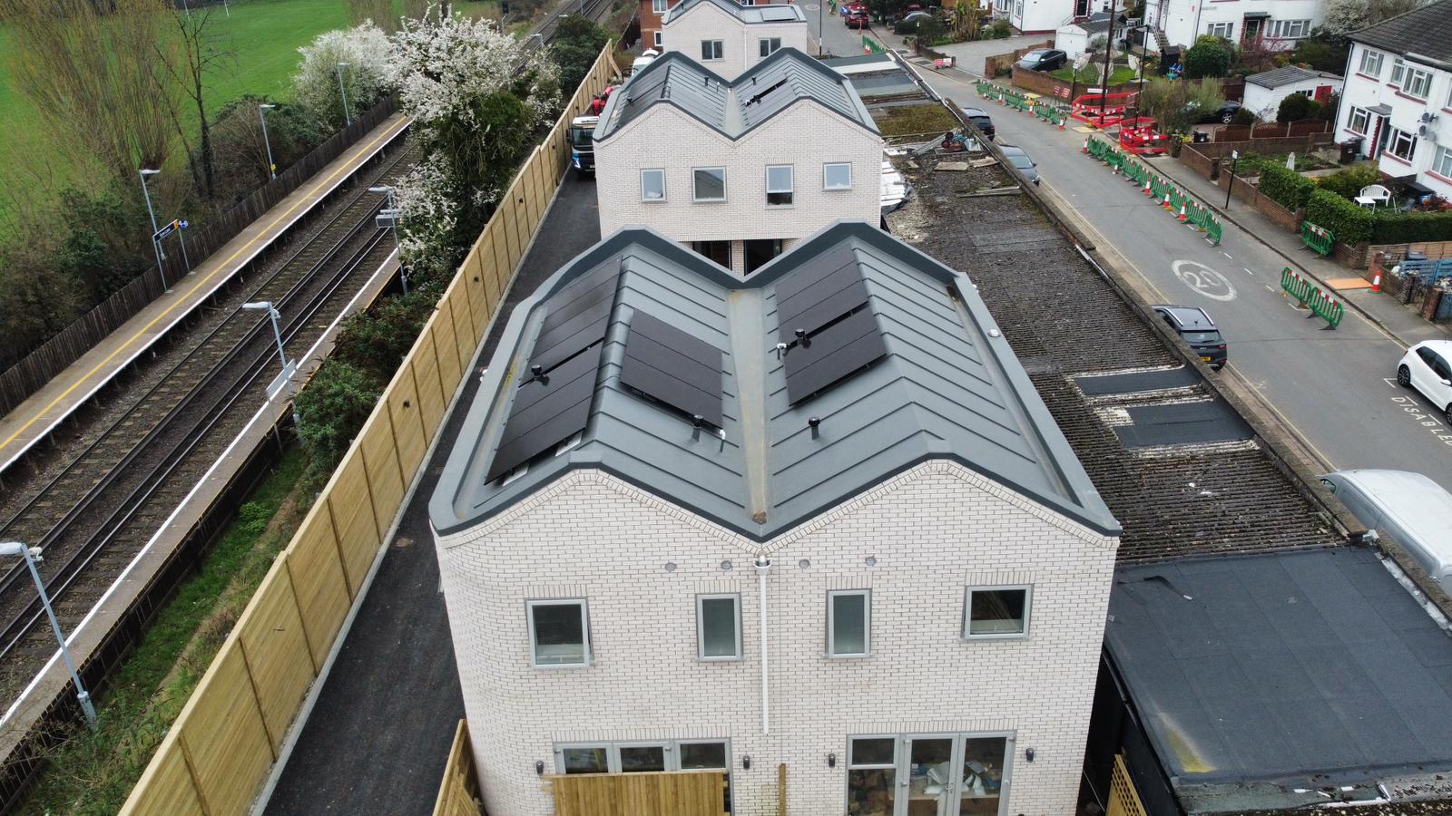 Aerial view of white residential building with distinctive dark grey pyramid-shaped roof, railway line visible to left, houses and road in background