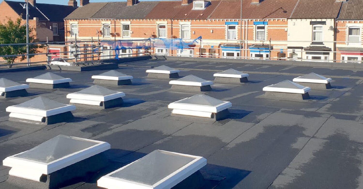 Flat rooftop with multiple white rectangular ventilation units arranged in rows, brick terraced houses visible beyond under blue sky.