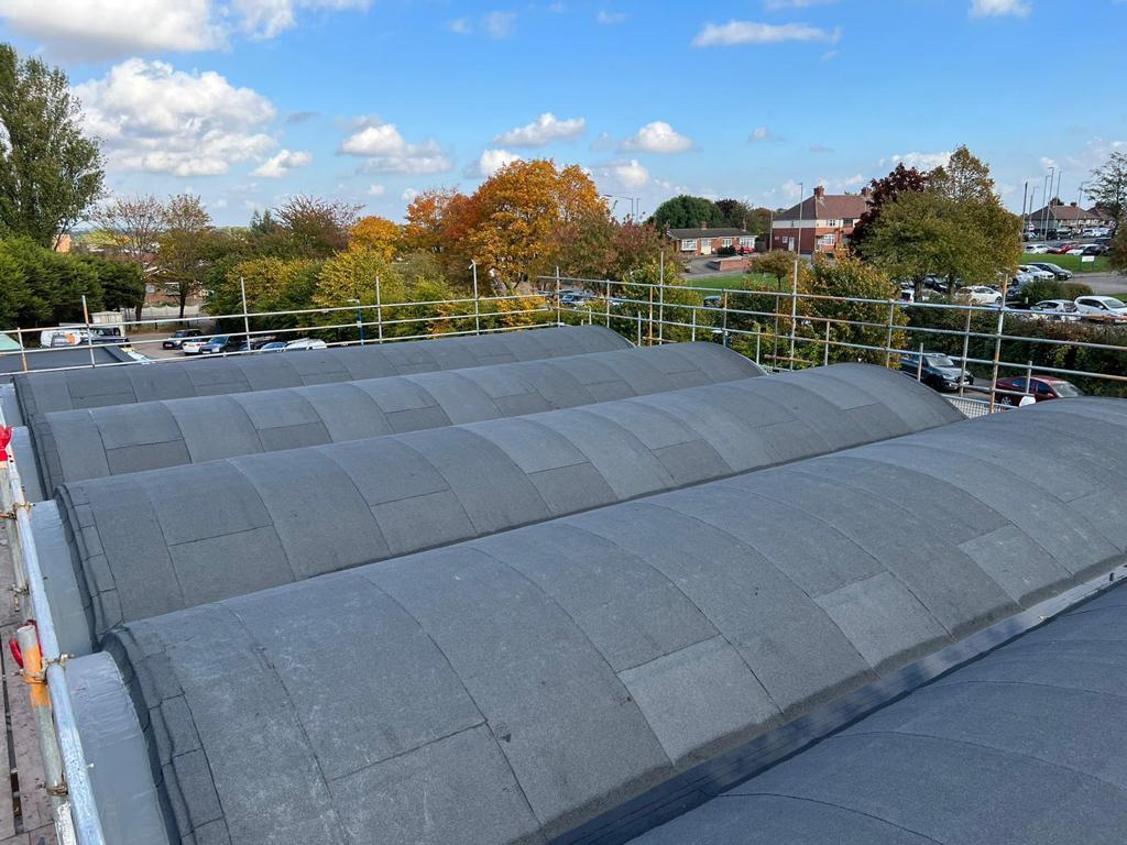 Flat roof with grey membrane covering, metal safety railings, autumn trees and residential buildings in background under blue sky.