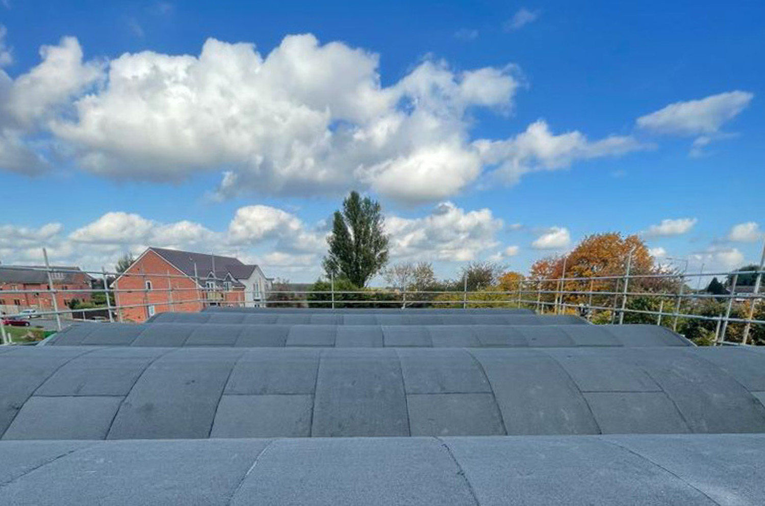 Large flat roof terrace with dark grey tiles, metal railings, and view of residential houses under blue sky with white clouds.