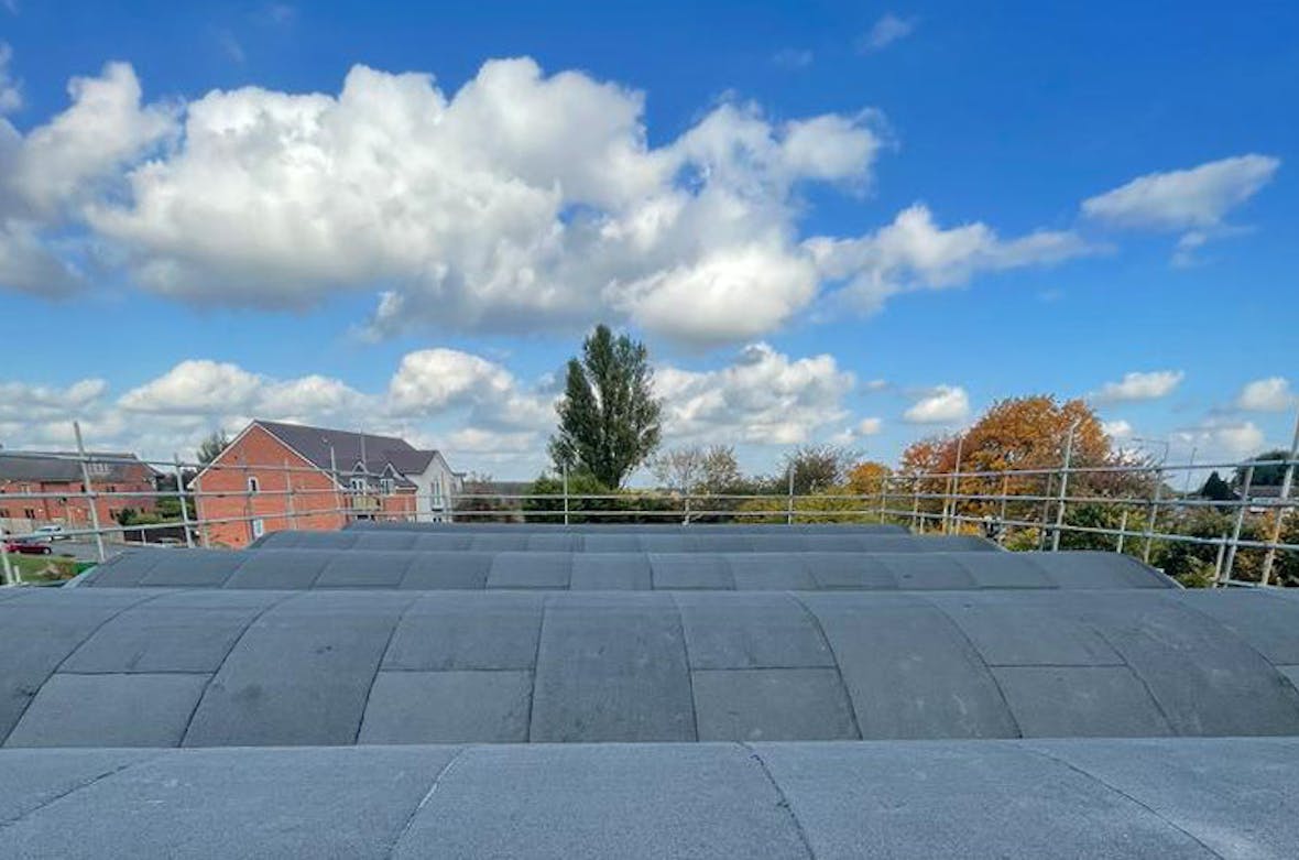 Grey tiled rooftop terrace with metal railings, residential houses and trees in background under blue sky with white clouds.