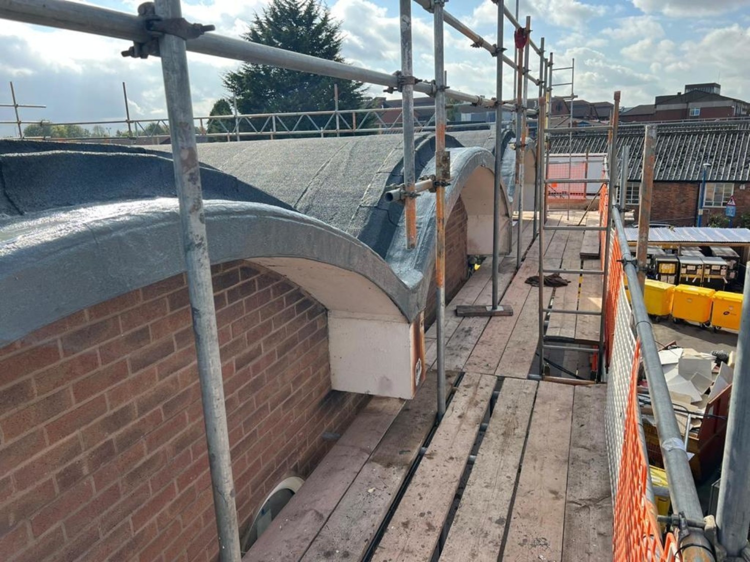 Construction site with metal scaffolding around curved brick and concrete structure, wooden planks, orange safety barriers.