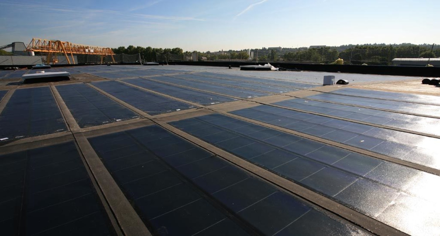Large solar panel installation on flat rooftop with blue panels arranged in geometric grid pattern, bridge visible in background.