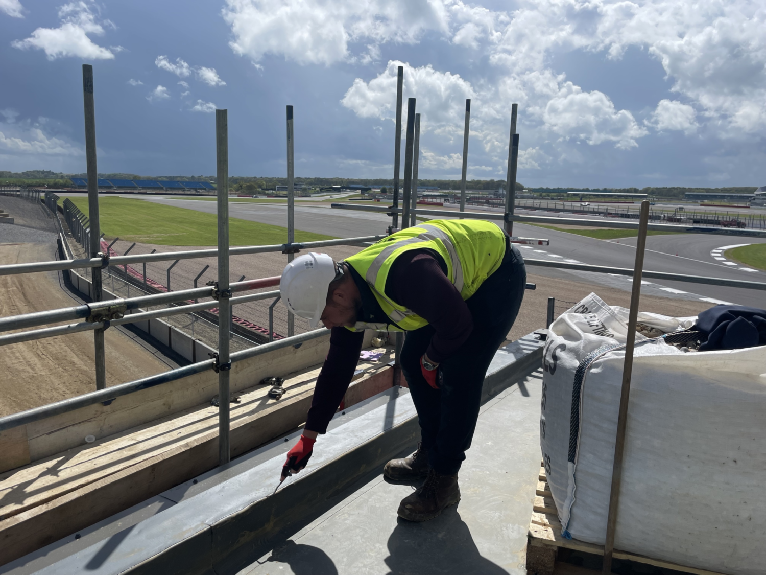 Worker in high-vis jacket on scaffolding platform working on rooftop with airport runway visible in background under cloudy sky.
