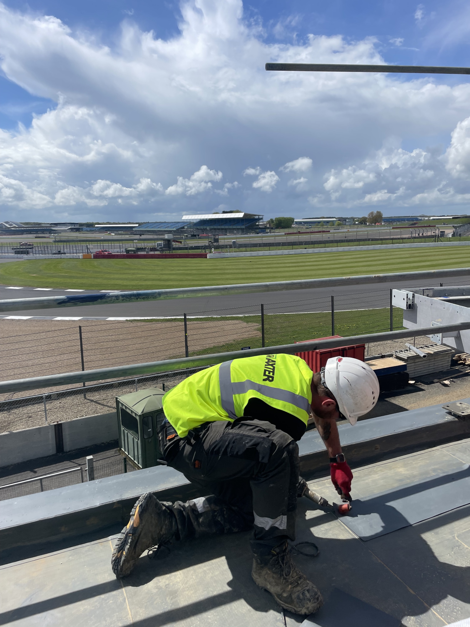 Worker in high-vis yellow jacket and white hard hat drilling on rooftop, with airport runway and terminal buildings visible in background.