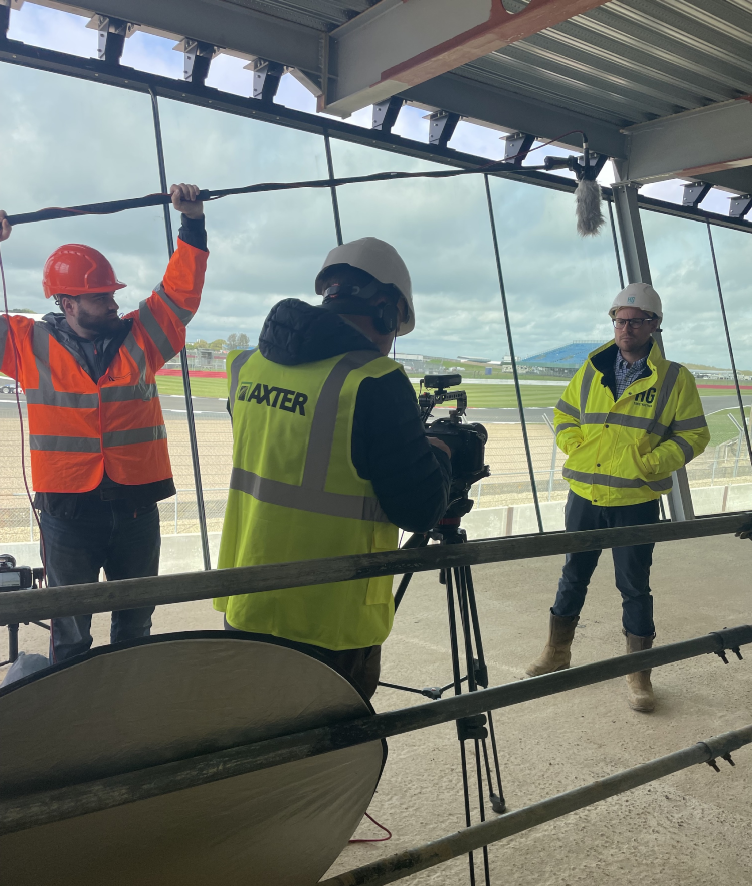 Three construction workers in hi-vis jackets and hard hats inside industrial building with large windows. Camera operator filming between them.