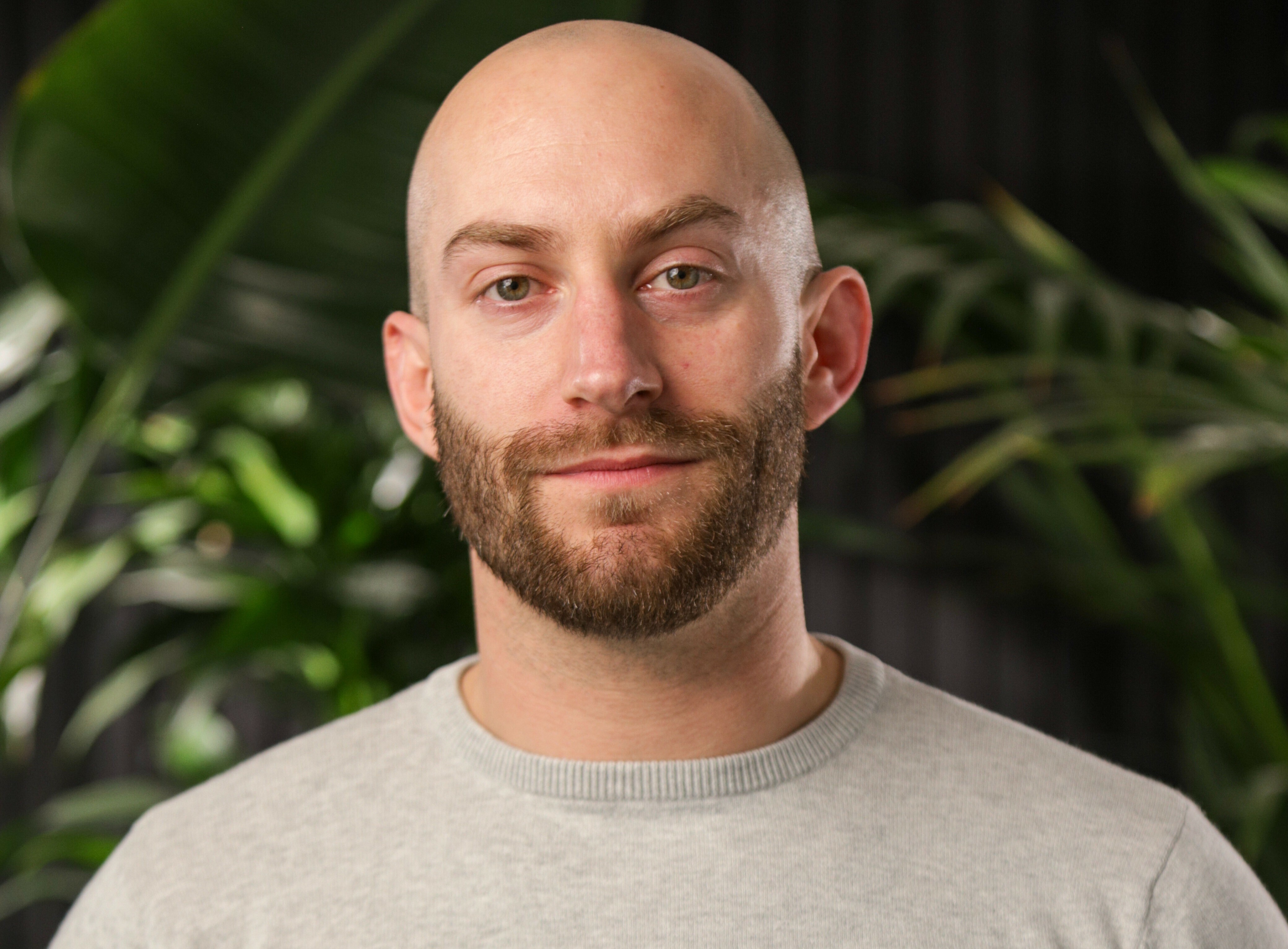 Bald man with brown beard wearing light grey jumper, green plants visible in blurred background.