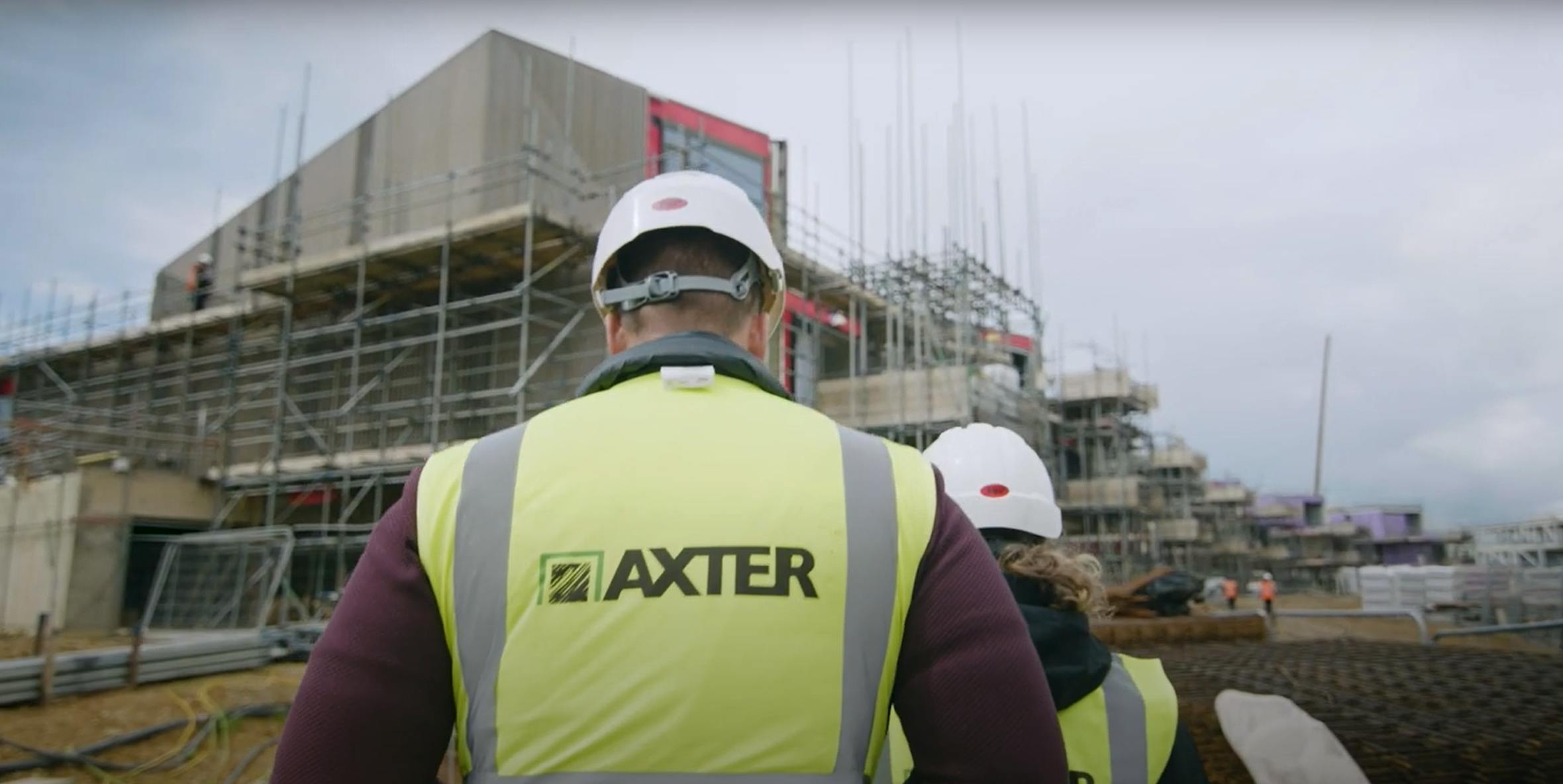 Construction worker in yellow high-vis vest with "AXTER" branding facing building site with scaffolding and concrete structures.