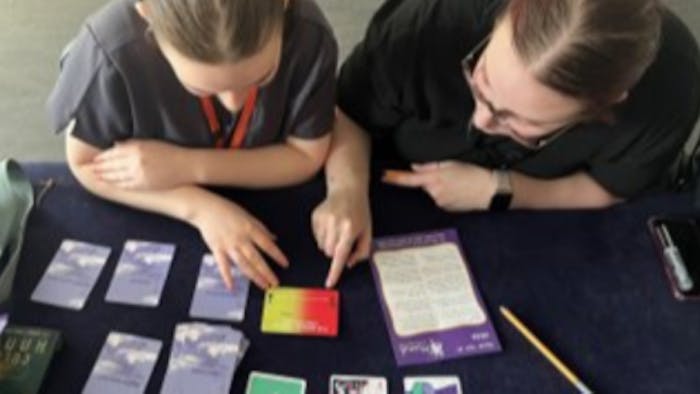 Two people in dark clothing examining coloured cards arranged on a dark surface, with one person holding a yellow and red card.
