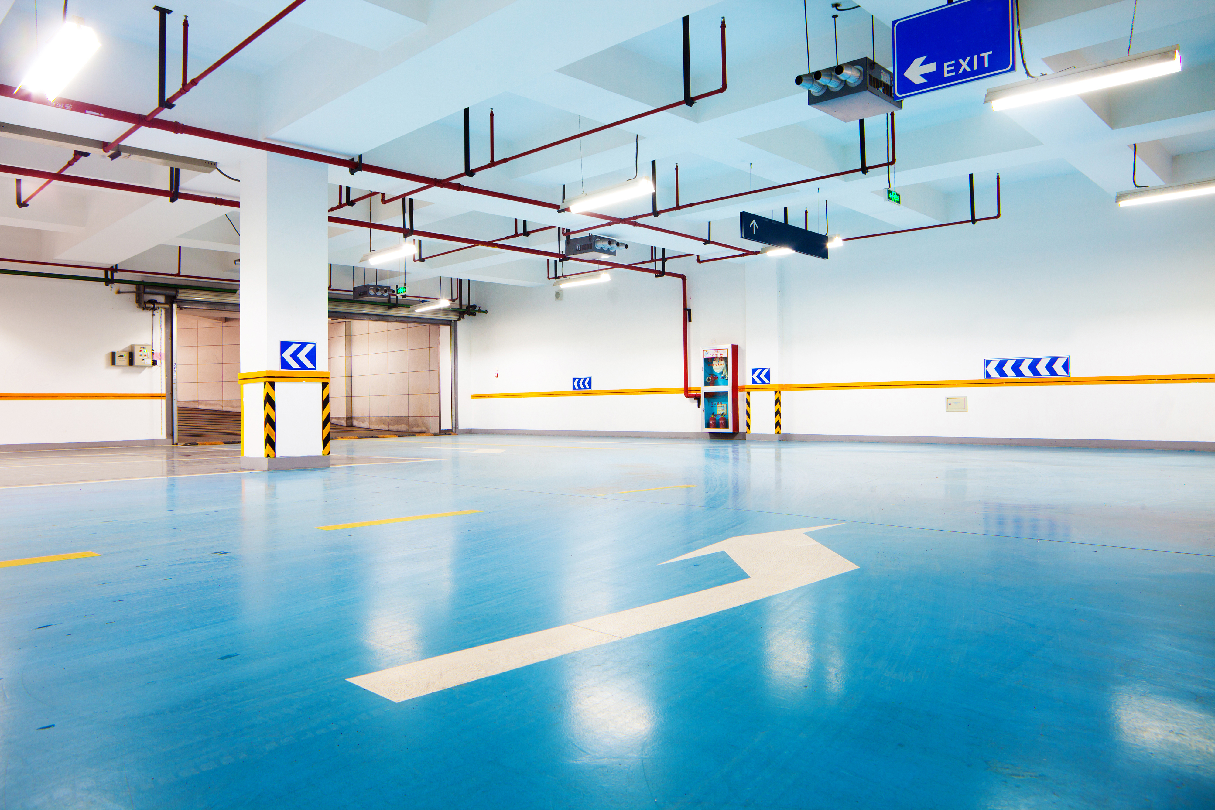 Underground car park with blue polished floor, white columns, red ceiling pipes, yellow striping, and blue exit signage.