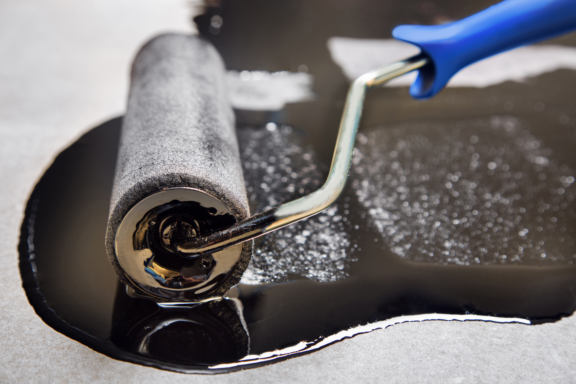 Person using paint roller with blue handle to apply grey paint, creating textured surface with visible paint splatter.
