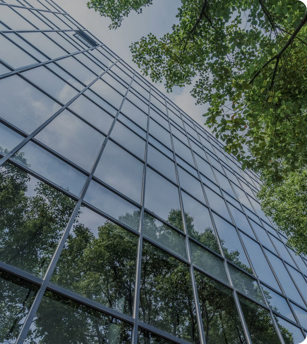 Low-angle view of glass office building with metal grid framework, green tree foliage visible on right side and reflected in windows.