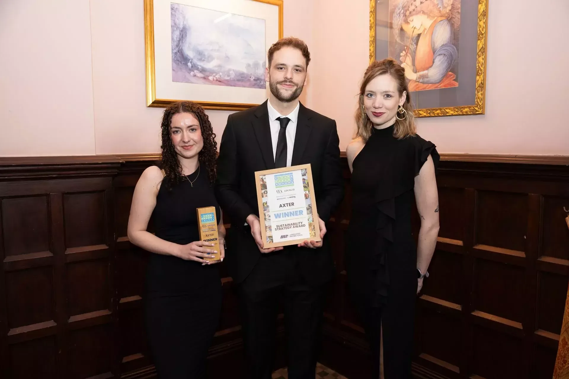 Three people in black formal wear holding awards and certificates in wood-panelled room with framed paintings on pink walls.