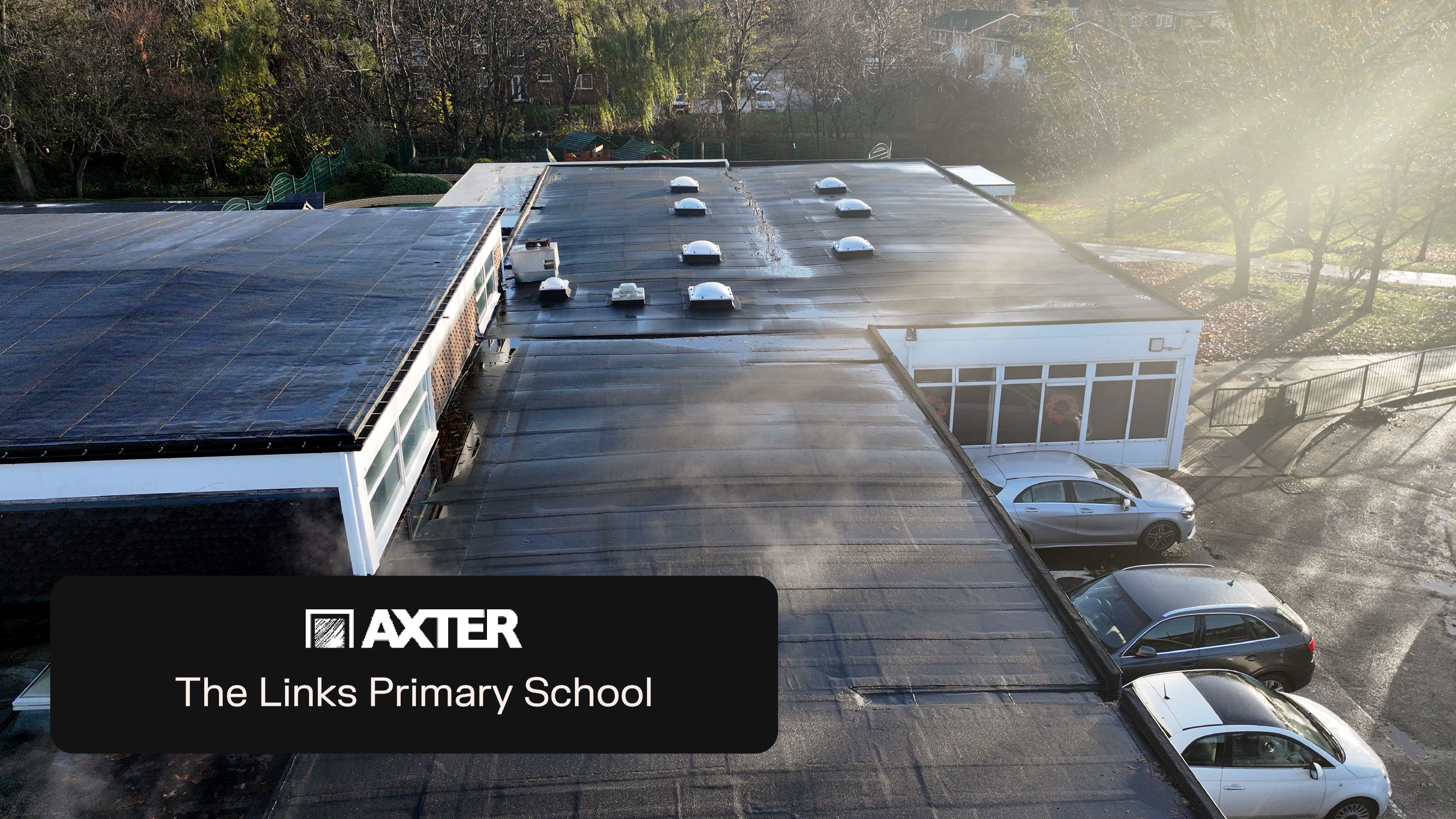 Aerial view of modern single-storey school building with flat roof, skylights, and car park. Trees visible in background.