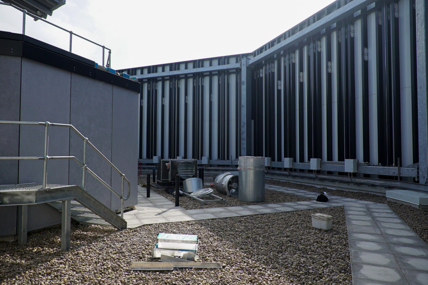 Modern concrete building courtyard with vertical metal cladding, external staircase, gravel ground, and overcast sky.