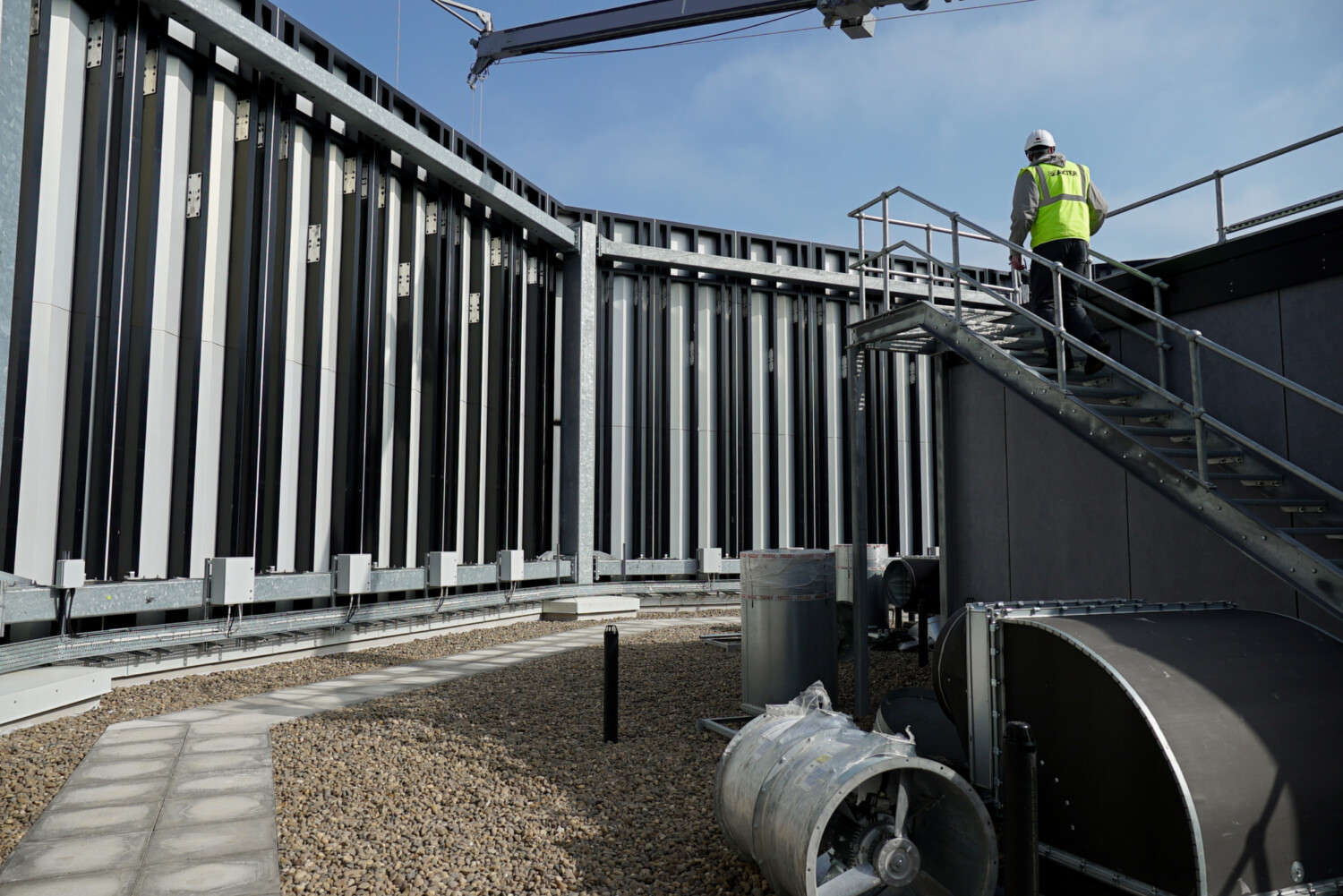 Construction worker in hi-vis jacket on metal staircase of industrial building with vertical steel cladding and curved pipe below.