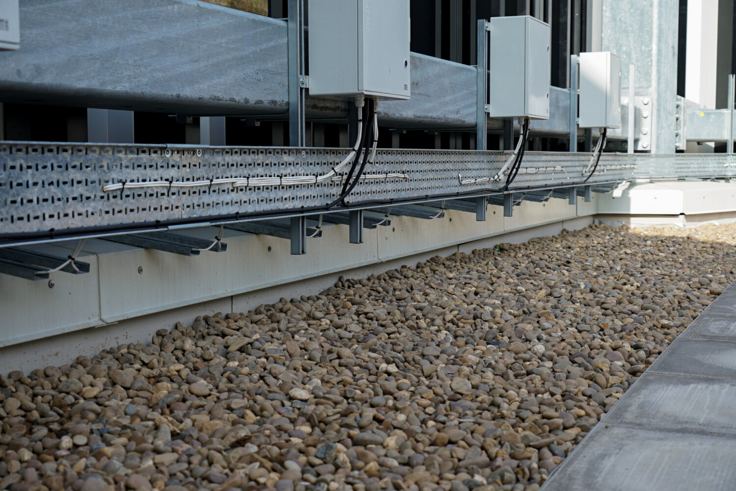 Metal structural framework with cables above brown gravel bed beside concrete pavement and white building exterior.