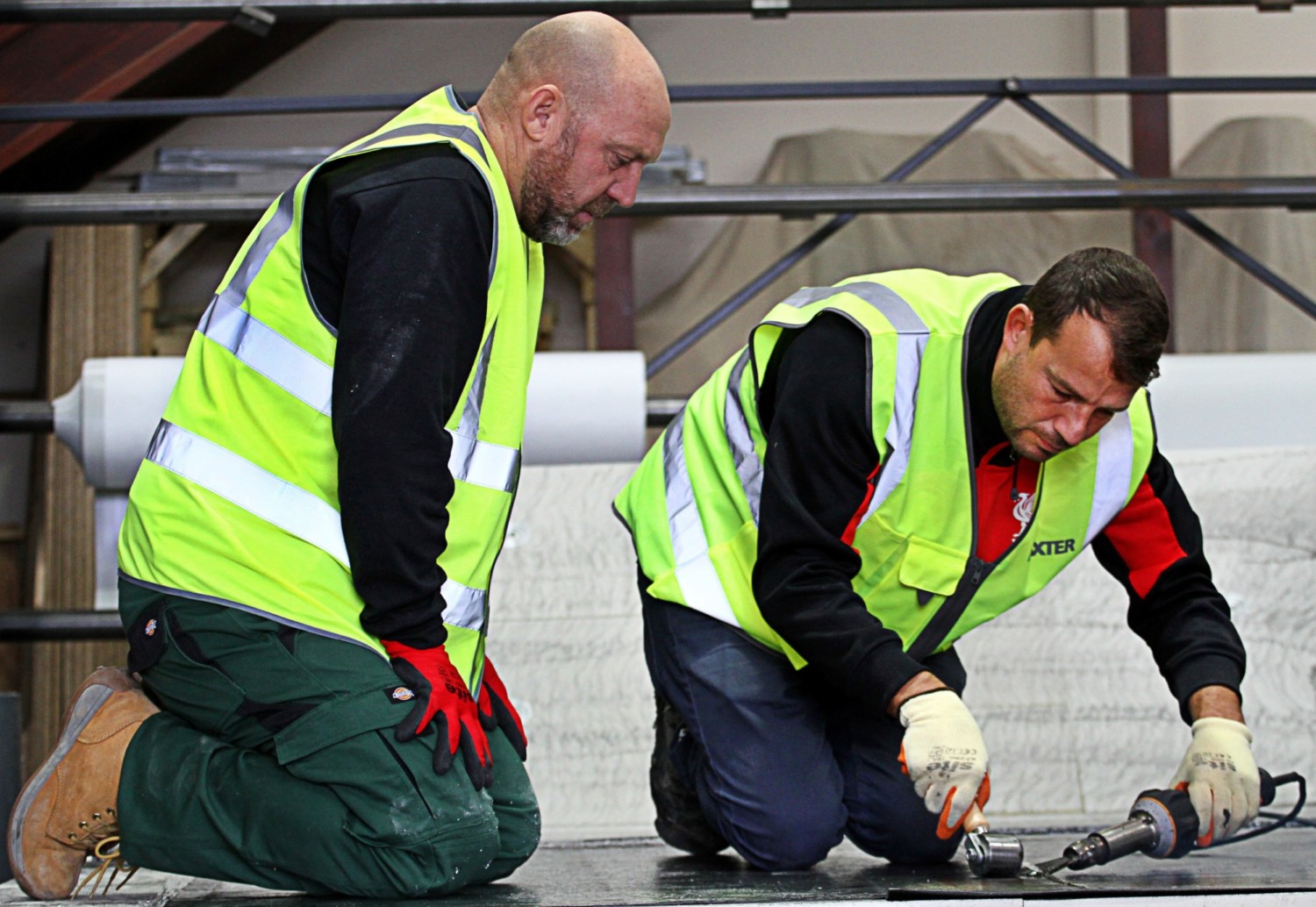 Two construction workers in high-vis yellow jackets kneeling on concrete, examining equipment with scaffolding in background.