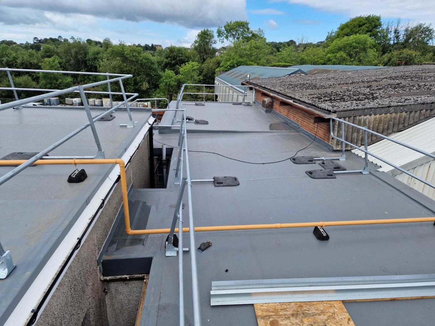 Grey flat roof with orange safety barriers, metal railings, and roof equipment. Adjacent buildings and green trees visible under cloudy sky.