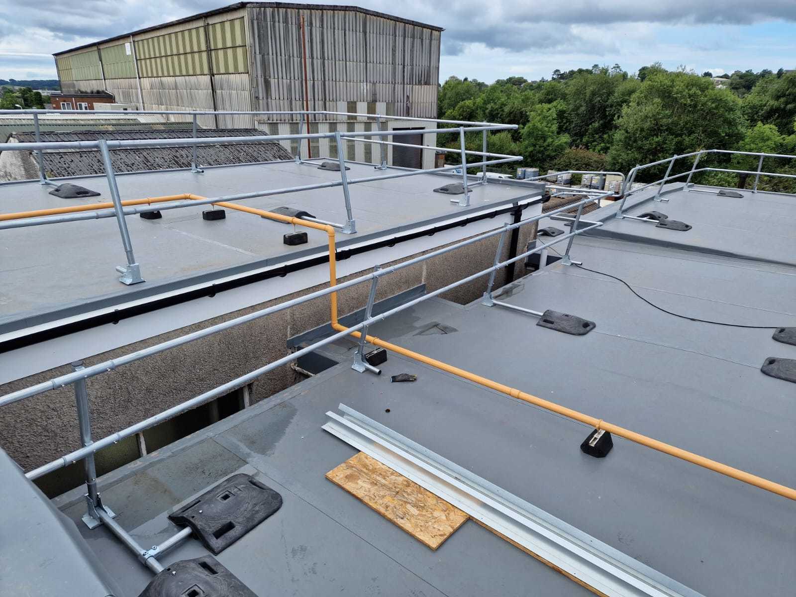 Grey flat roof with metal safety railings, orange cable, and brown wooden planks. Large corrugated warehouse visible in background.