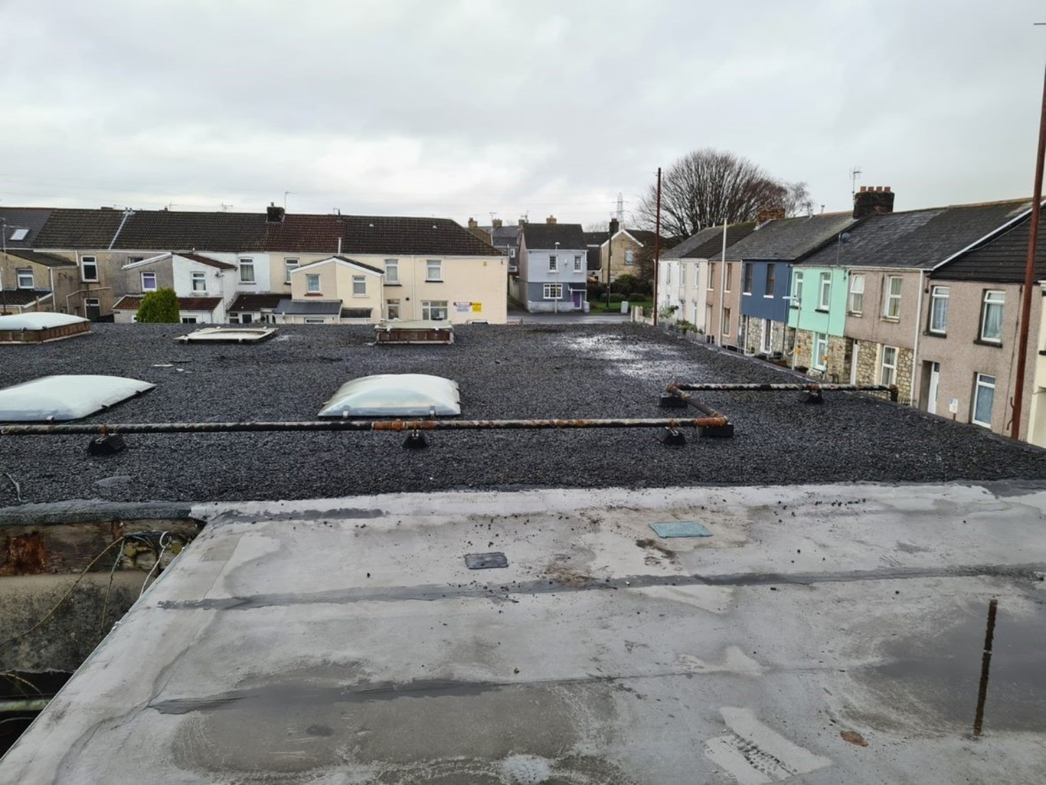 Flat roof with gravel surface and skylights, overlooking terraced houses with coloured facades under overcast sky.