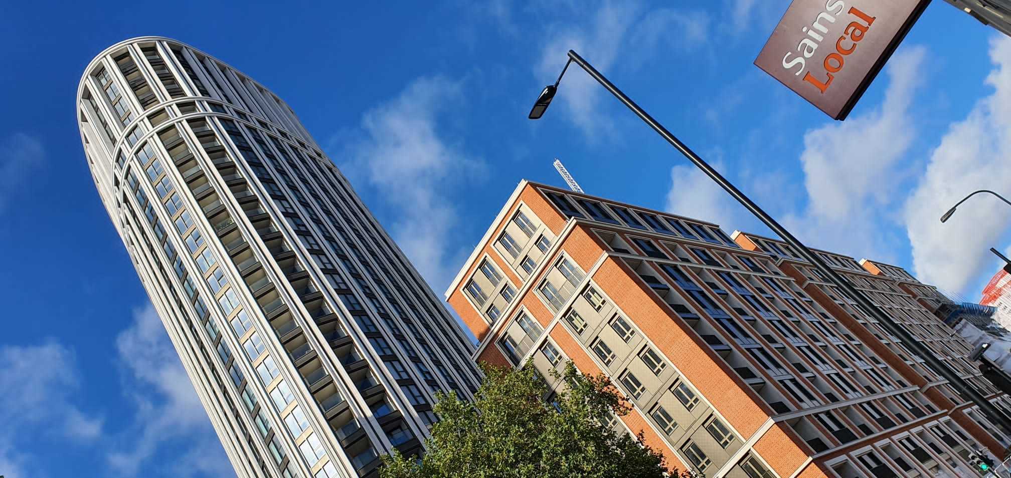 Tall cylindrical glass tower and orange brick residential building against blue sky with white clouds, viewed from below.
