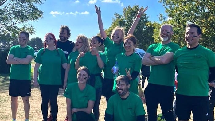 Group of people in matching green shirts posing outdoors with arms raised, trees and blue sky in background.