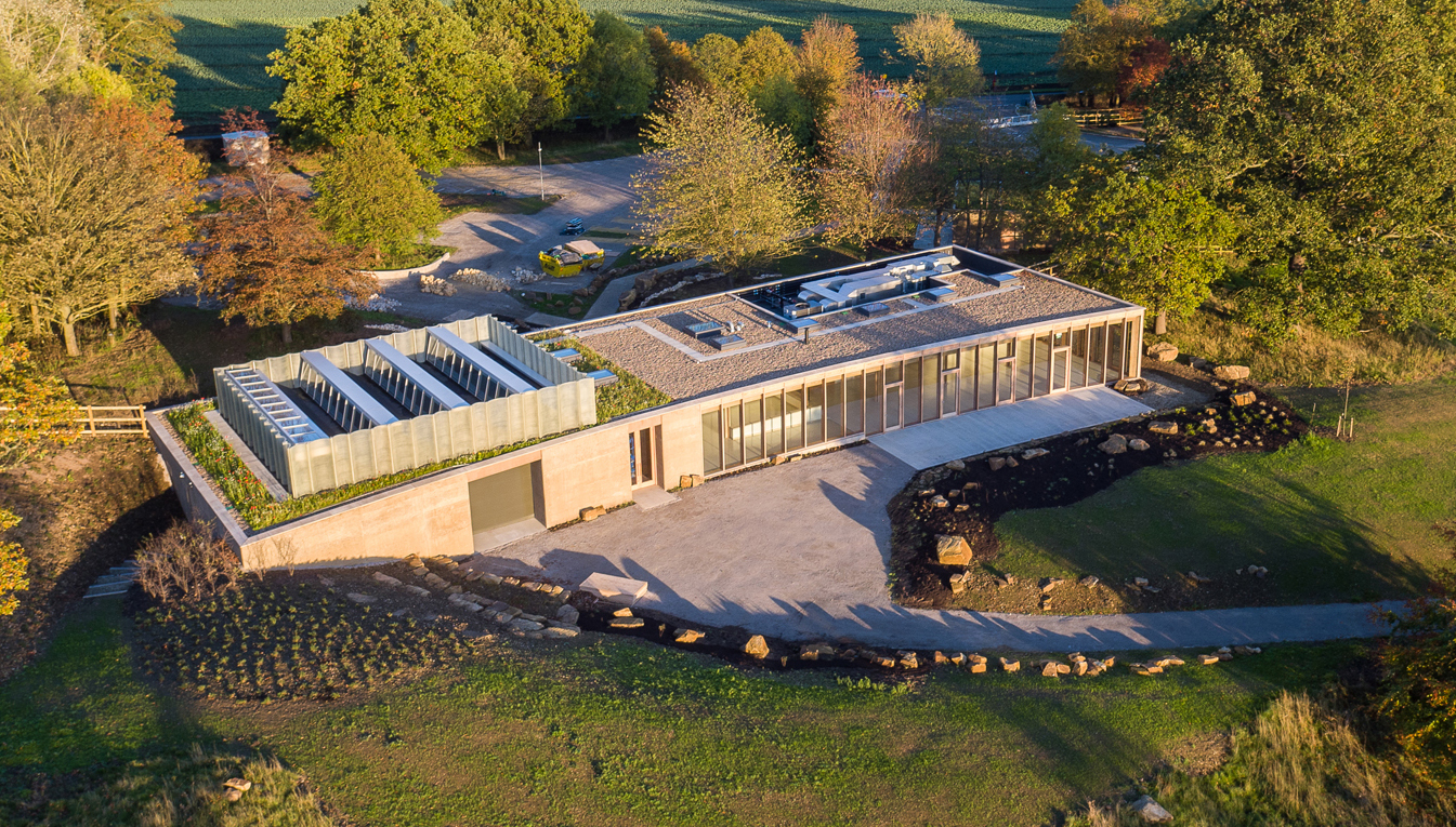 Modern single-storey building with flat roof and glass walls, surrounded by autumn trees and green fields, curved driveway visible.