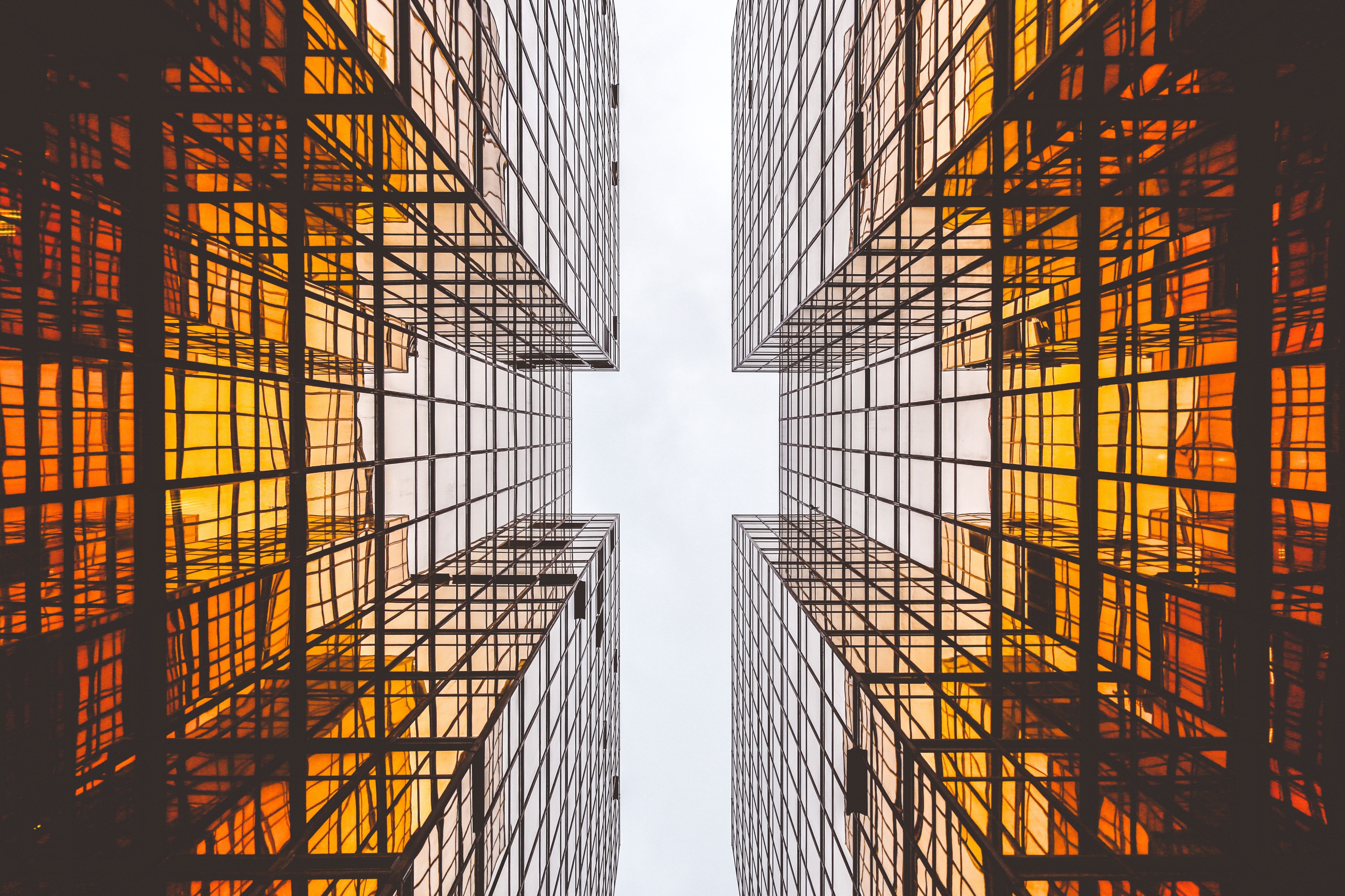 Upward view between tall glass buildings with orange and yellow reflections creating geometric patterns against grey sky.