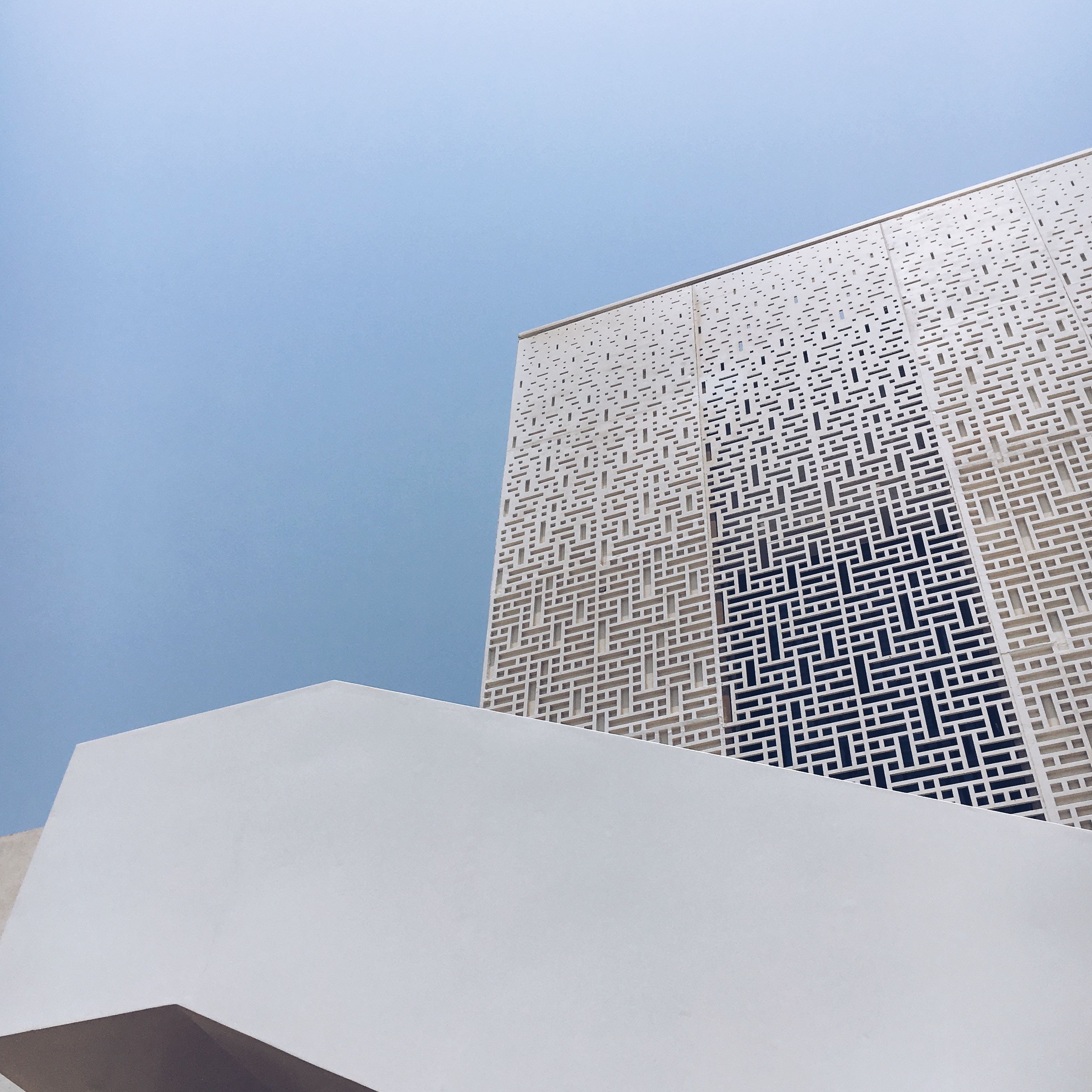 Modern white building with geometric black pattern on upper facade against pale blue sky, curved white structure below.