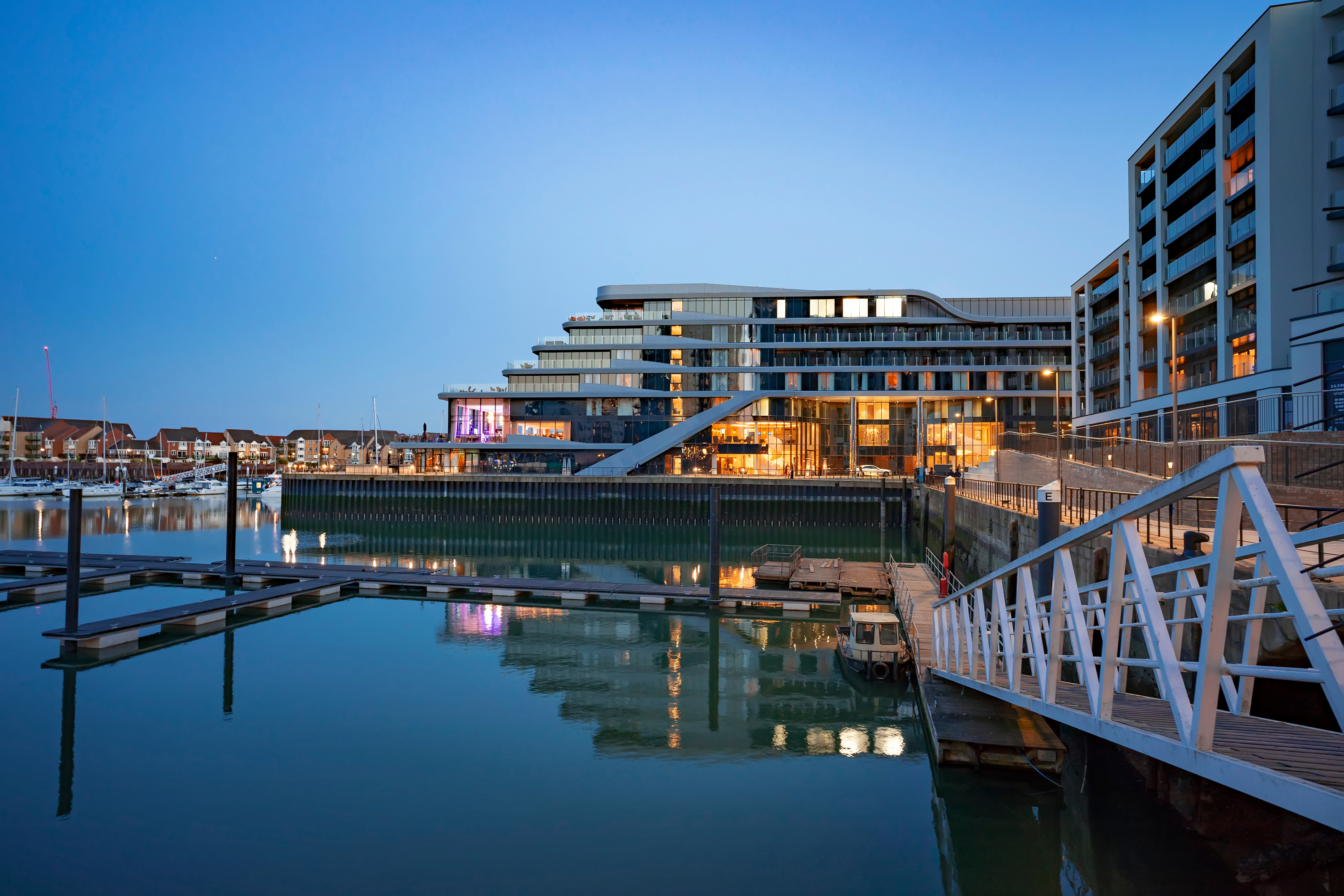 Modern waterfront buildings with illuminated windows reflected in calm harbour water, white wooden boardwalk in foreground, dusk sky.