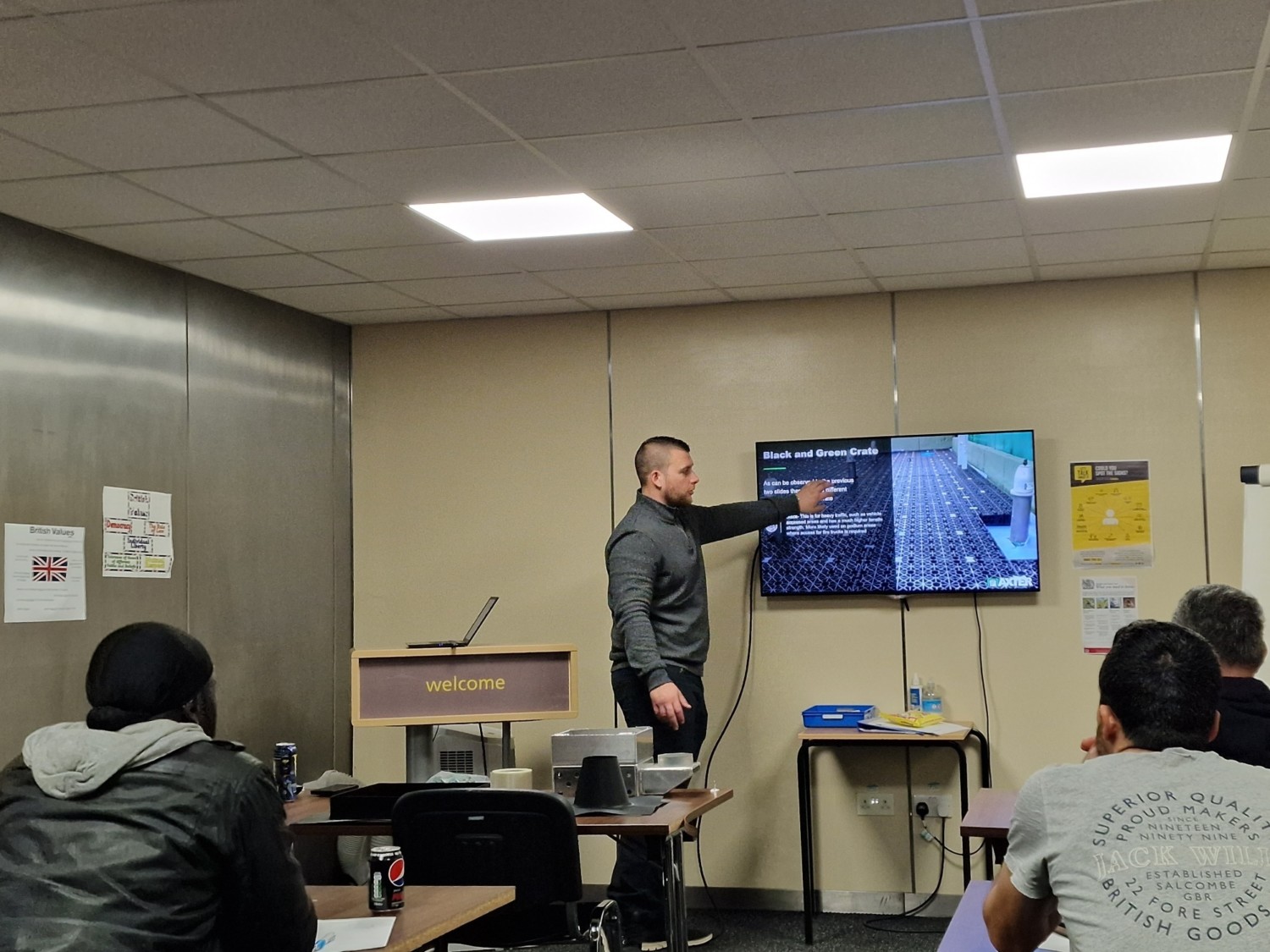 Man in dark shirt pointing at wall-mounted screen in classroom with seated students at desks under fluorescent lighting.