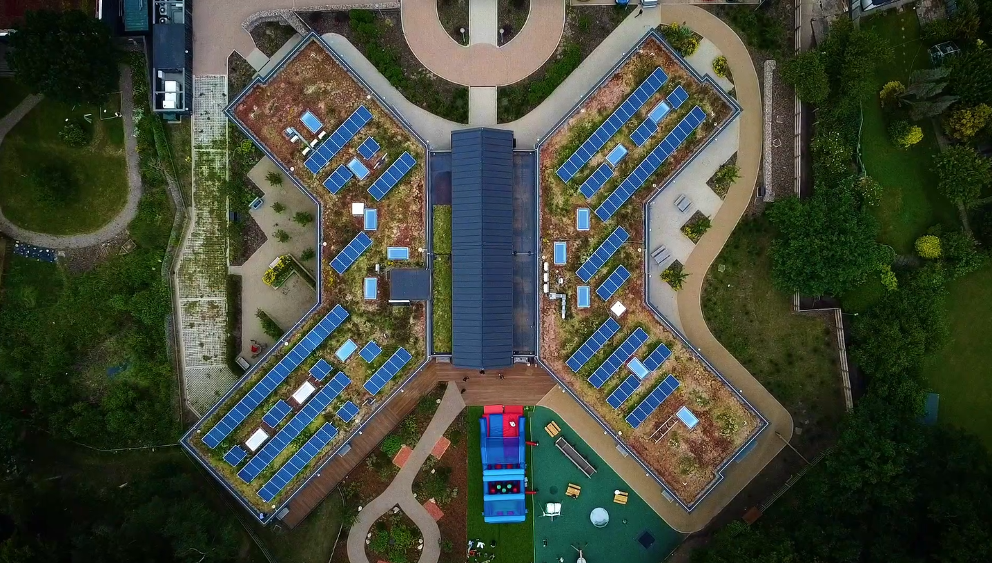 Aerial view of a cross-shaped building with green roofs covered in blue solar panels, surrounded by trees and pathways.