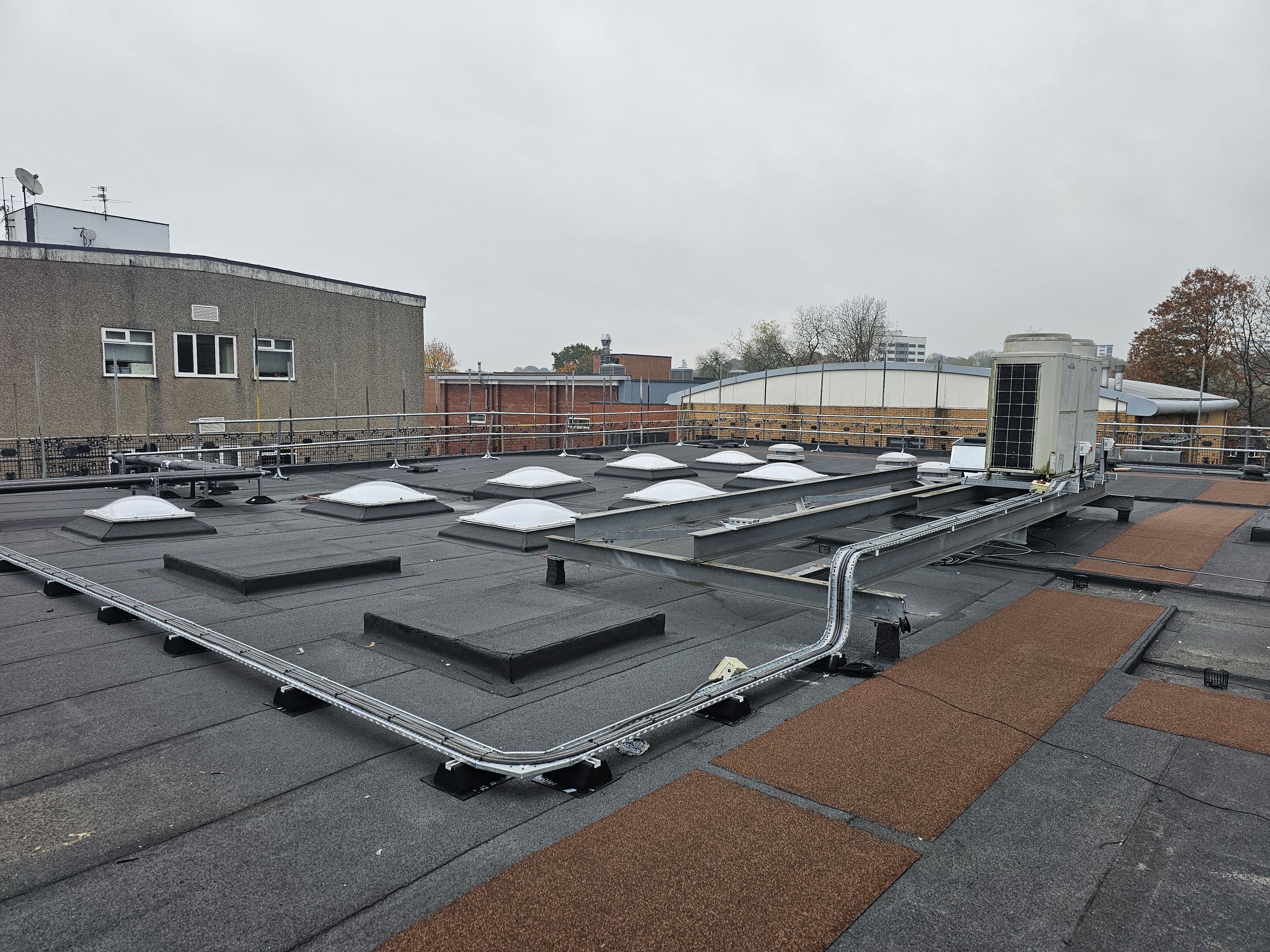 Flat roof with multiple rectangular grey ventilation units, metal pipework, and orange-brown weatherproofing sections under overcast sky.